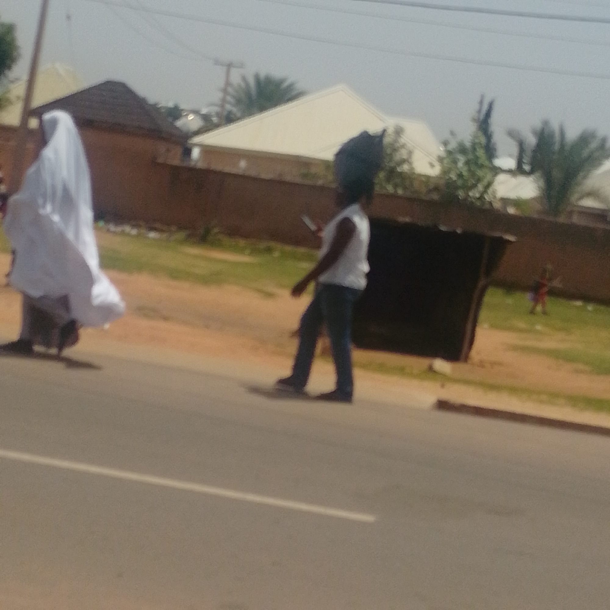 In Gombe, Nigeria, a woman in a white hijab walks on a dusty road. She is accompanied by another person wearing a dark head covering and a white top, holding a mobile phone. In the background, a partially visible open-air structure with a corrugated roof stands, and behind it, there are residential buildings and vegetation. A child in a red and blue outfit is seen in the distance on the grassy area. The scene suggests a daytime setting with a bright sky.