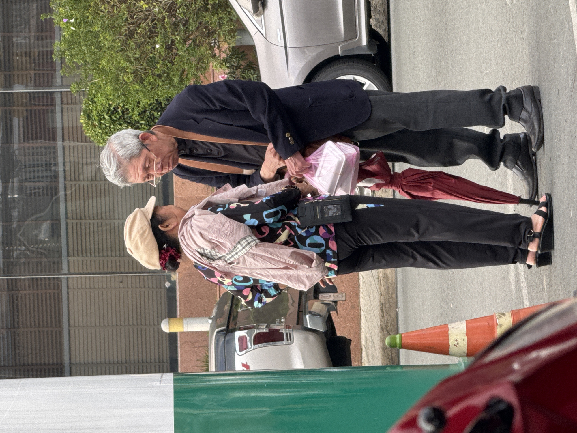 Two individuals, a man and a woman, stand on a street in Da Lat, Vietnam, appearing to be in conversation. The man, wearing glasses, a dark jacket, and dark pants, holds a bag of food items in his right hand. The woman, in a tan cap and a colorful jacket, faces the man, with her left hand touching his right. A red umbrella is partially visible near their feet. In the background, a silver car and a pickup truck are parked, with a green pole and an orange traffic cone in the foreground. The setting suggests an urban environment.