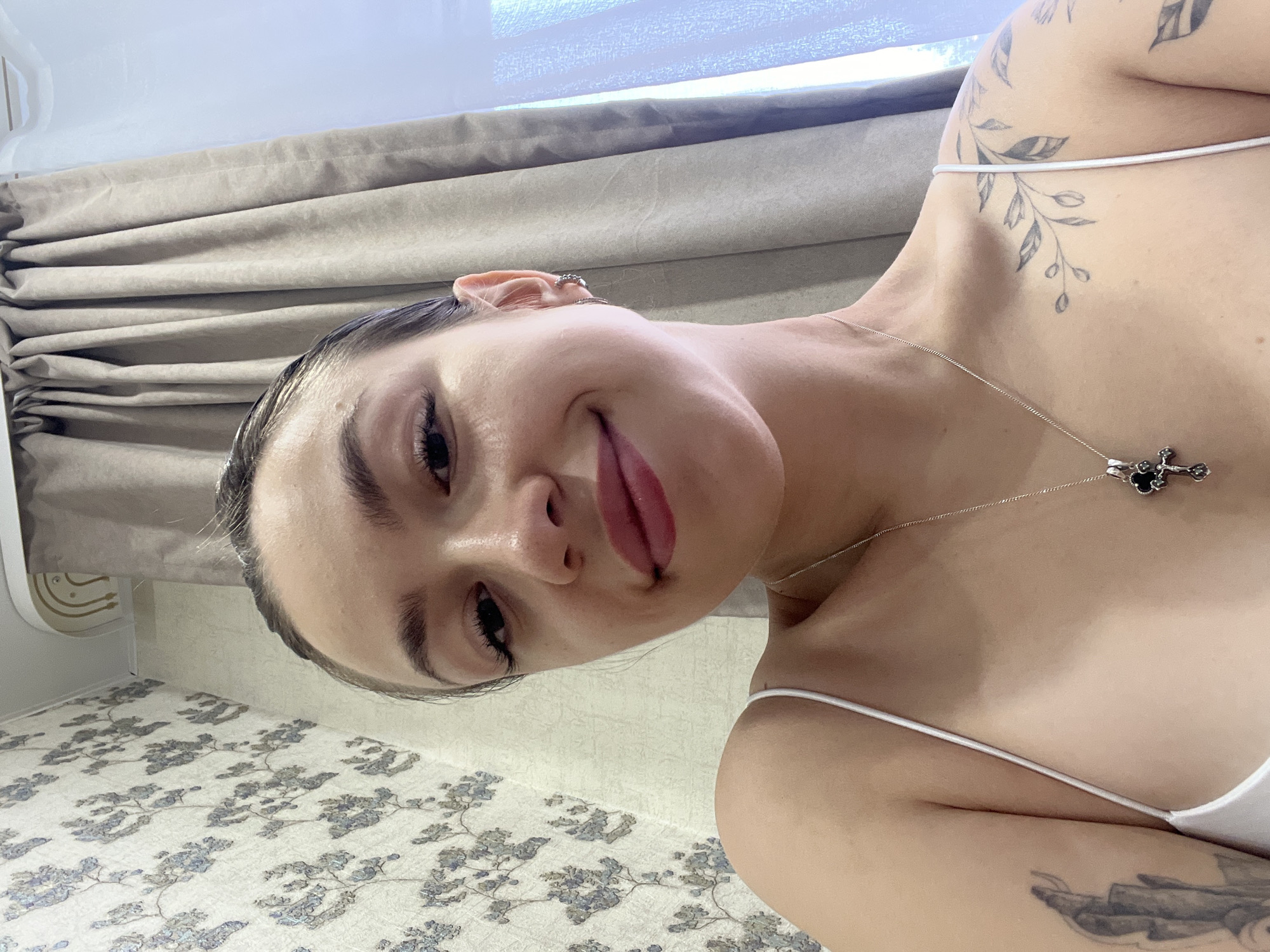 A young woman with dark hair pulled back is taking a selfie indoors. She is smiling slightly and wearing a white camisole and a silver necklace with a cross pendant. She has tattoos on her left arm and shoulder. The background shows a wall with floral wallpaper and a beige curtain. This image was taken in Chaikovsky, Russia.