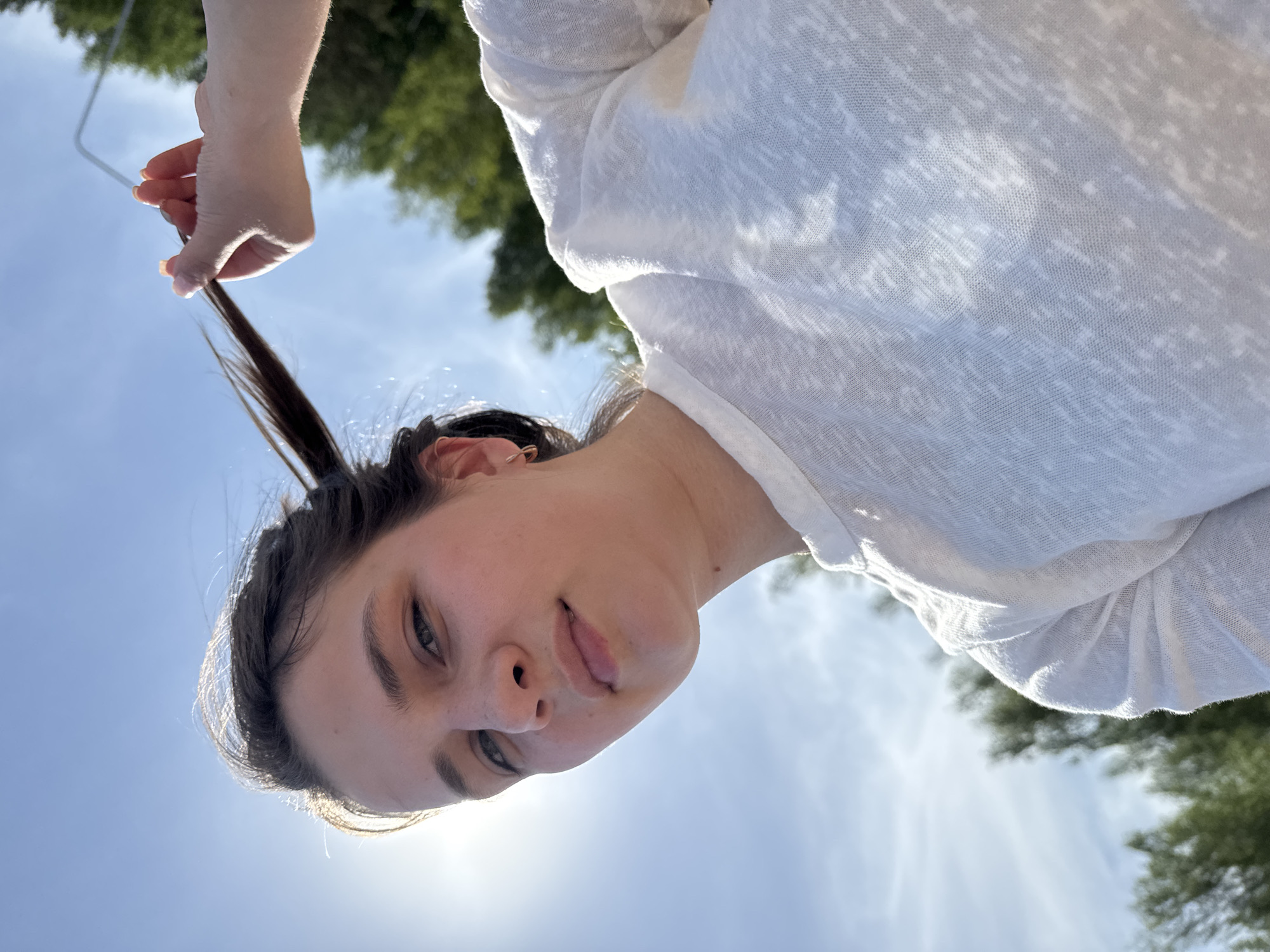 A fair-skinned person wearing a white, long-sleeved shirt is holding a strand of dark hair. The setting appears to be outdoors, with a blue sky and some trees visible in the background. The image is oriented with the person's face towards the bottom. Based on the prompt, this location is in the city of Chaikovsky, Russia.