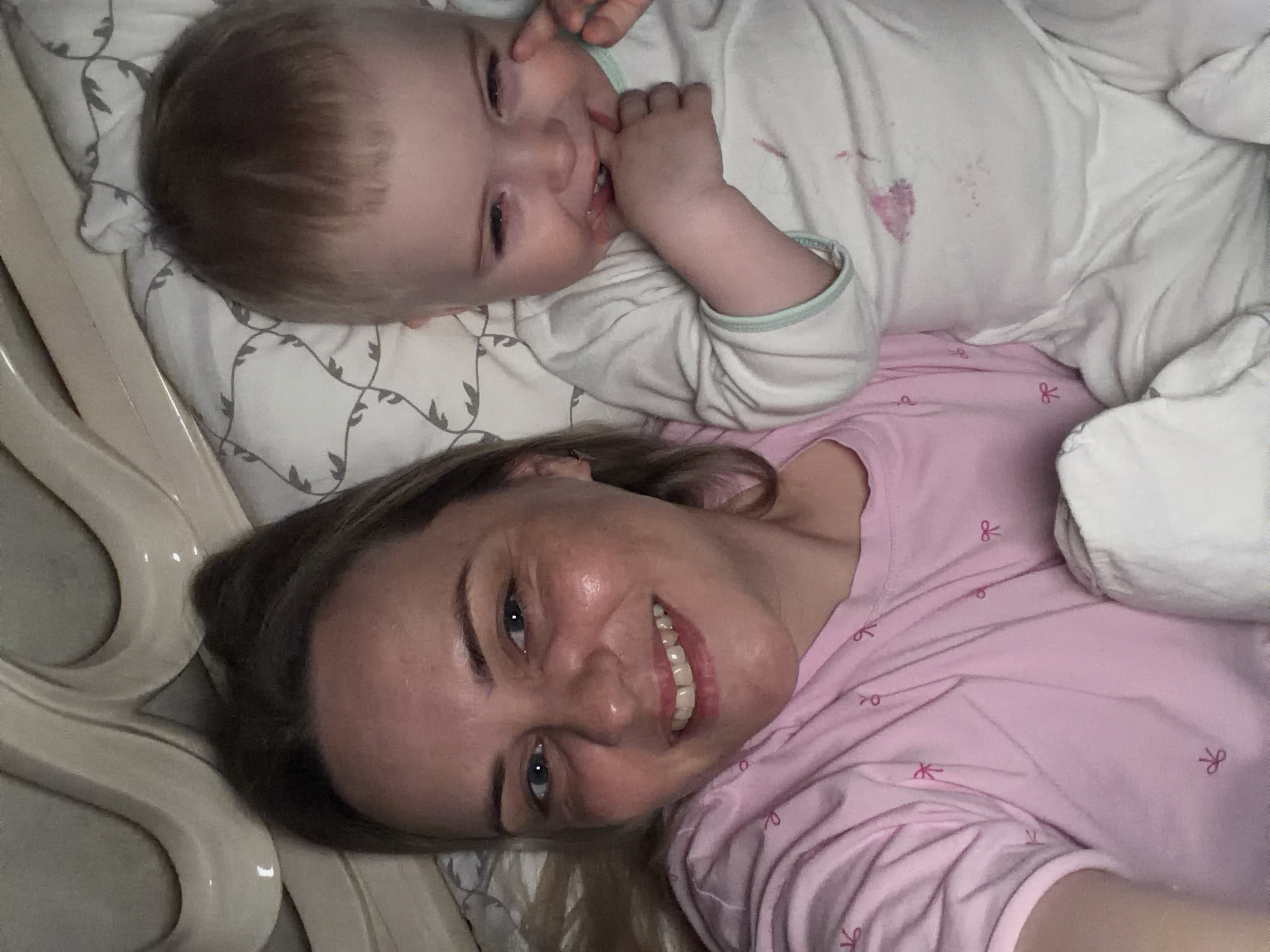 A woman and a baby are lying on a bed. The woman, wearing a pink shirt with a bow pattern, is positioned at the bottom of the frame, smiling at the camera. The baby, dressed in white pajamas, is lying above her, with a hand near their mouth. They are both lying on a patterned sheet, and part of a white blanket is visible on the right. The environment is a bedroom, likely a private residence. There is no information available in the image to indicate that the location is in Votkinsk, Russia.