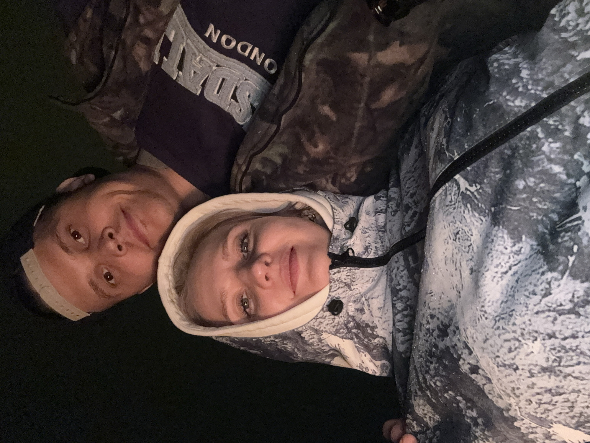 Two individuals are in the image. A man with a baseball cap and a dark t-shirt with "NDON" printed on it is to the left. A woman with blond hair wearing a hooded jacket is to the right. They are posing close together. The environment appears dark. No definitive elements suggest a location in Mayak, Russia.