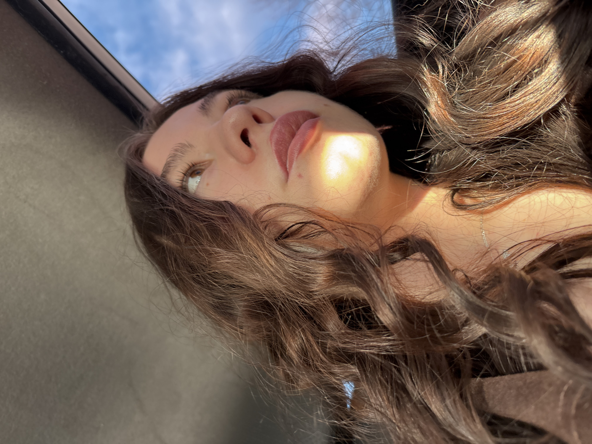 A close-up, angled portrait features a young woman with long, wavy, dark brown hair, illuminated by natural sunlight, in Perm, Russia. Her head is tilted, and she is looking upwards, revealing the underside of her chin and jawline. Her left eye is visible, with light brown irises and long eyelashes. Her lips are full with a natural pinkish hue. Strong sunlight falls on her face, creating a bright highlight on her right cheek and upper lip, and catching the strands of her hair, making them glisten with lighter brown and golden tones.
The background shifts from a clear blue sky with white clouds in the upper left, suggesting an outdoor or partially open-air setting, to a darker, possibly interior, space towards the bottom and right. A dark, thin structure, possibly a window frame or car roofline, is visible along the top edge of the image. The woman's dark clothing is subtly visible on her shoulder, and a delicate silver necklace can be seen around her neck, partially obscured by her hair. Her expression is calm and thoughtful as she gazes upwards. There is no visible text in the image. The time of day is clearly daytime, given the bright sunlight and blue sky.