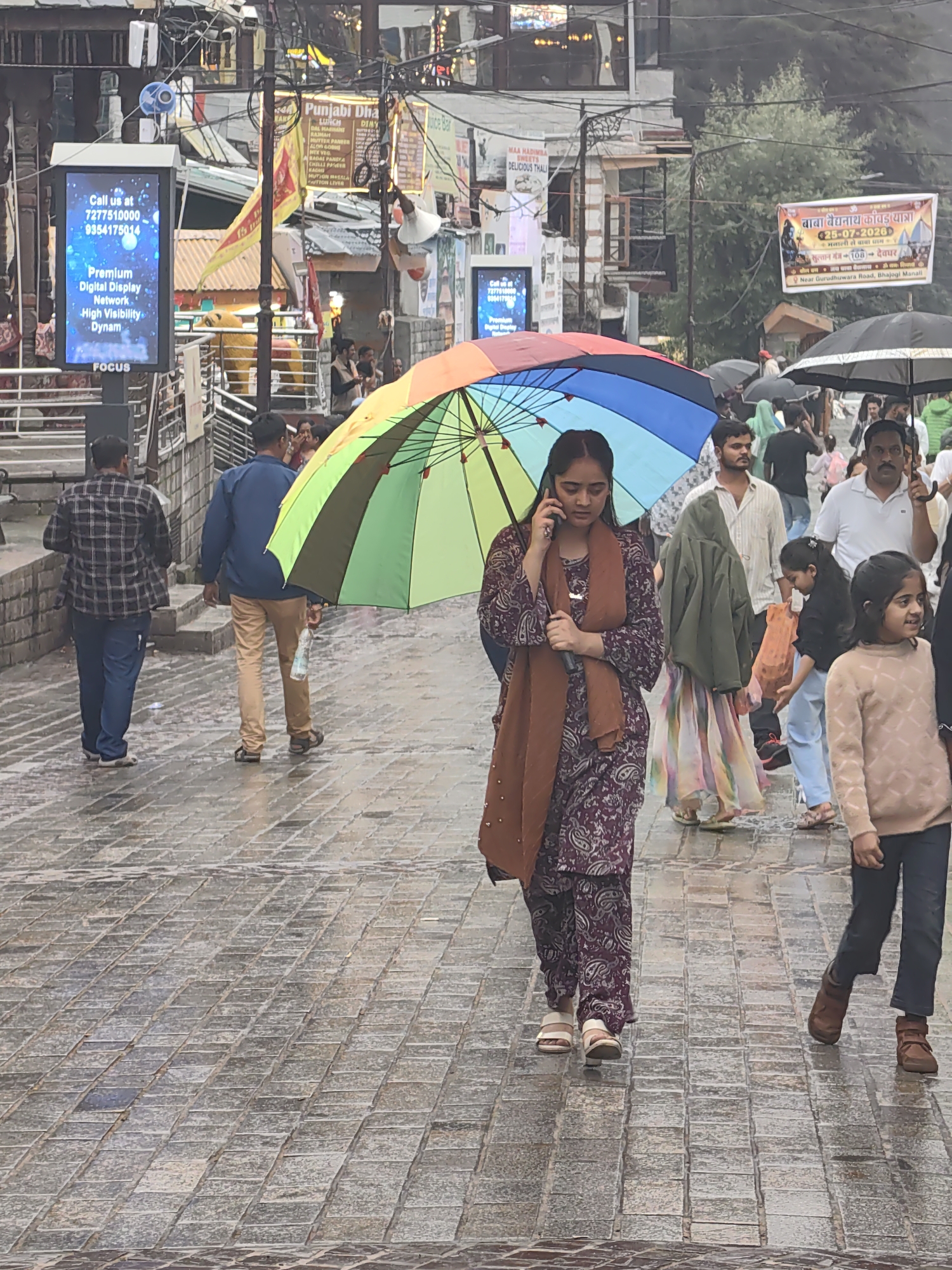 The image depicts a street scene in Manali, India, likely a busy tourist area, on a rainy day. The ground is wet and reflects the ambient light, suggesting recent rainfall. Several people are visible, many holding umbrellas. In the foreground, a woman is walking while talking on her cell phone, holding a colorful rainbow umbrella. She is wearing a patterned shalwar kameez and a brown scarf. To her right, a young girl is also walking, holding an orange bag. Further back, other individuals are seen, some with umbrellas, navigating the wet street. In the background, there are shops and signs, including a digital billboard advertising "Premium Digital Display" with contact information. The overall atmosphere is that of a public space experiencing inclement weather.