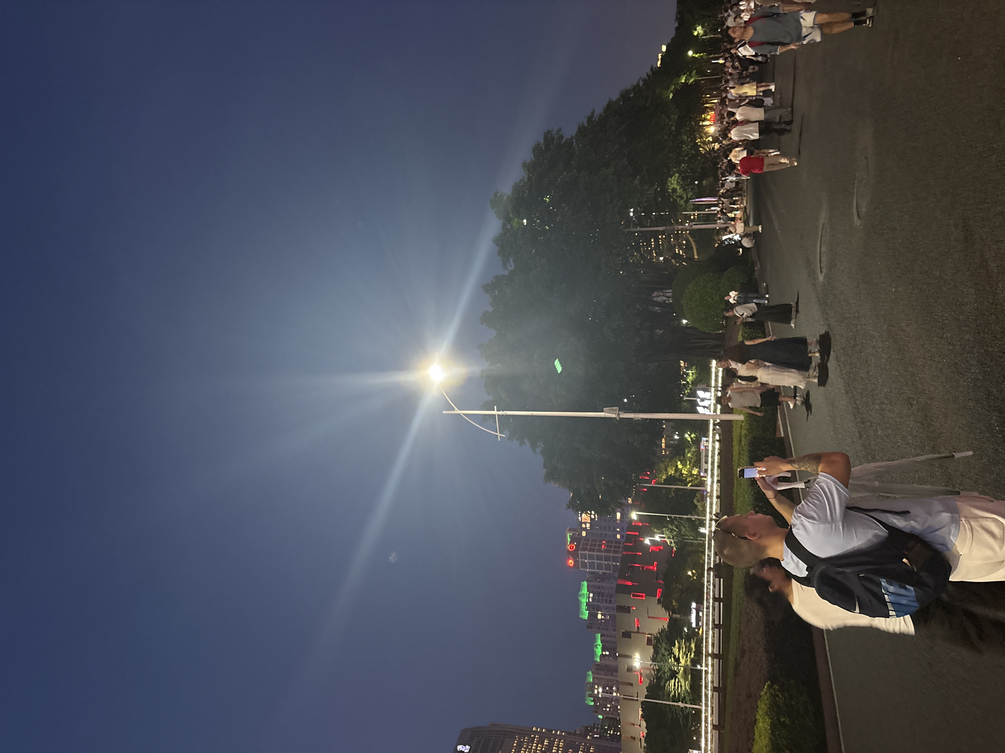 In Tianhe District, China, a wide asphalt road is illuminated by a bright streetlamp with a distinct light flare reaching across the dark blue twilight sky. A lush, large tree with dark green foliage stands prominently in the middle ground, its branches obscuring the view of distant city buildings. These buildings are lit with various colors, including green and red lights on their upper floors, suggesting a vibrant urban environment.
A group of people are walking along the road, some appearing to be tourists. In the foreground, on the left side of the image, a man with short blonde hair and sunglasses on his head is holding up a smartphone, appearing to take a photo. He is wearing a light blue t-shirt, white pants, and a dark backpack with blue accents. Beside him, a person in a white t-shirt and dark pants is partially visible. Other individuals are scattered across the road, some walking towards the viewer and others away, engaged in various activities. The scene suggests a casual evening gathering or stroll in a public urban space.