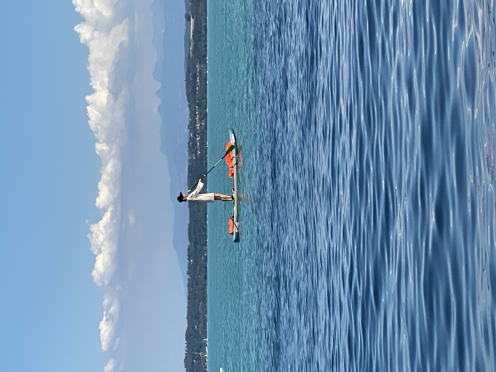 A person is stand-up paddleboarding on a body of water. The person is wearing a light-colored shirt and a dark baseball cap, and they are holding a paddle. The paddleboard has two orange life vests secured to it. The water is calm with ripples, and there is a distant shoreline with trees and buildings. Mountains are visible in the background, and the sky is blue with scattered white clouds. The scene appears to be a recreational activity on a lake.