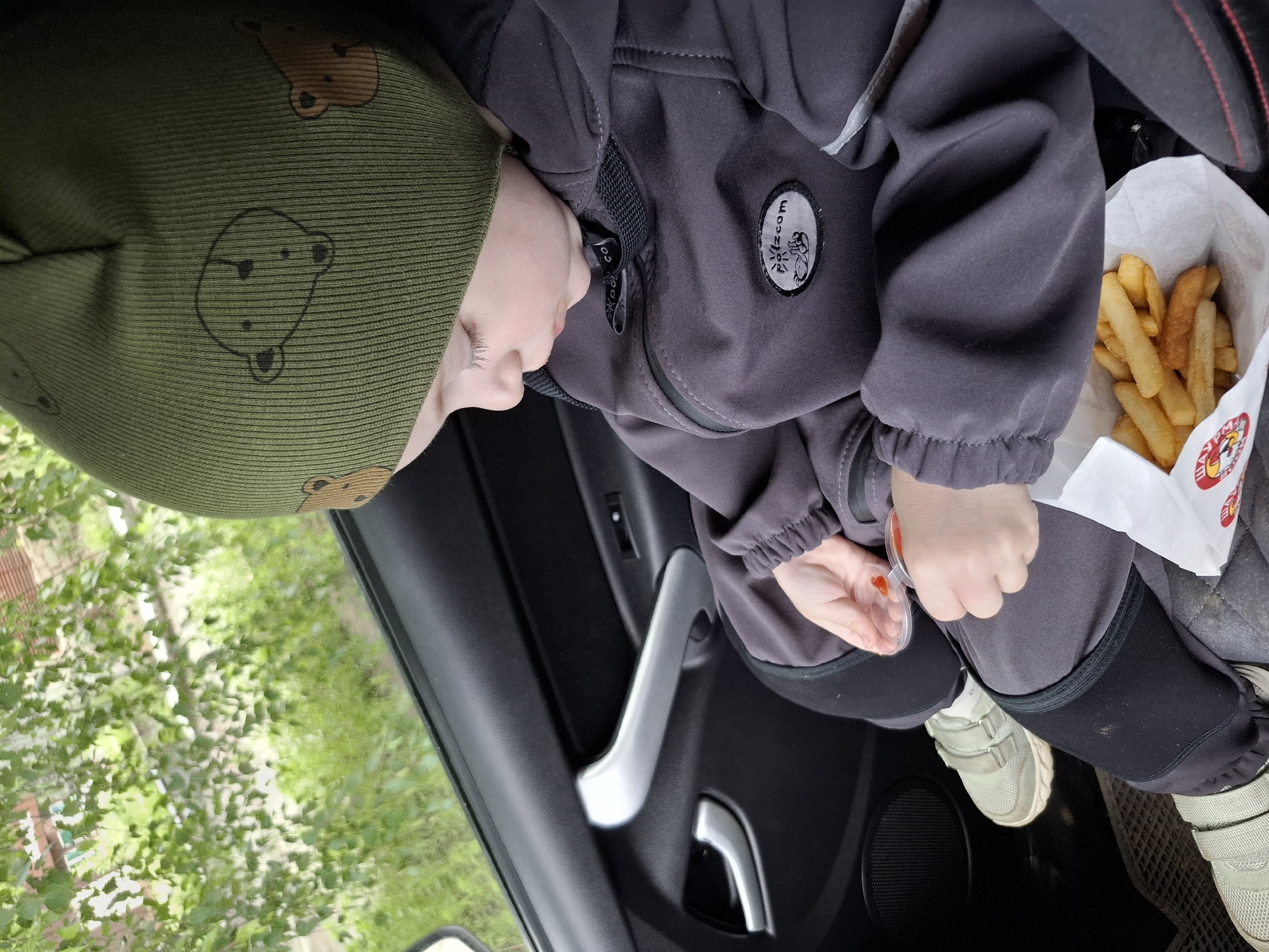 A young child is seated in the rear passenger compartment of a car in the city of Chaikovsky, Russia. The child, with fair skin and light eyelashes, wears an olive green ribbed knit beanie patterned with outline drawings of brown bears, a dark grey softshell jacket with a "Polizcom" brand patch on the left chest and a reflective stripe on the right sleeve, dark grey pants with black knee patches, and light green sneakers with three Velcro straps. The child holds two clear plastic portion cups containing red ketchup, looking down intently. A white paper bag with Cyrillic text "ШАУРМА" (SHAURMA) and "ЧАЙКОВСКИЙ" (CHAIKOVSKY) and a chicken head logo, containing golden-brown French fries, rests near the child's lap. The car's interior is visible, featuring a dark grey door panel with silver handles, a power window switch, and a black speaker grille. Outside the car window, blurred green foliage, indicative of trees and grass, is visible under daylight conditions.