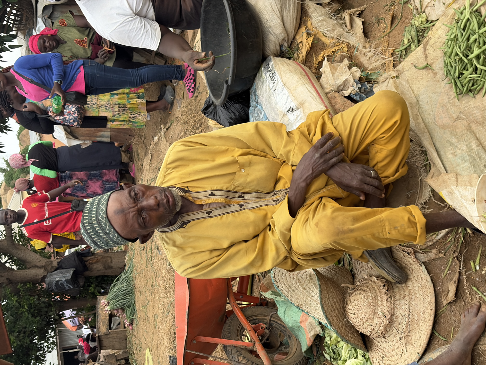The image depicts an outdoor market scene in Gura Top, Nigeria. In the foreground, an elderly man wearing a yellow traditional outfit and a green patterned cap is seated on the ground. He has a weathered face and his hands are resting on his lap. Next to him, there are two woven straw hats and a pile of green beans. In the background, several people are visible, some appearing to be vendors and shoppers. A red wheelbarrow is situated to the left of the seated man, and various sacks and bags are scattered around the market. The ground is sandy and dry, and trees are visible in the distance, suggesting a rural or semi-rural setting. The overall atmosphere appears to be a bustling marketplace.