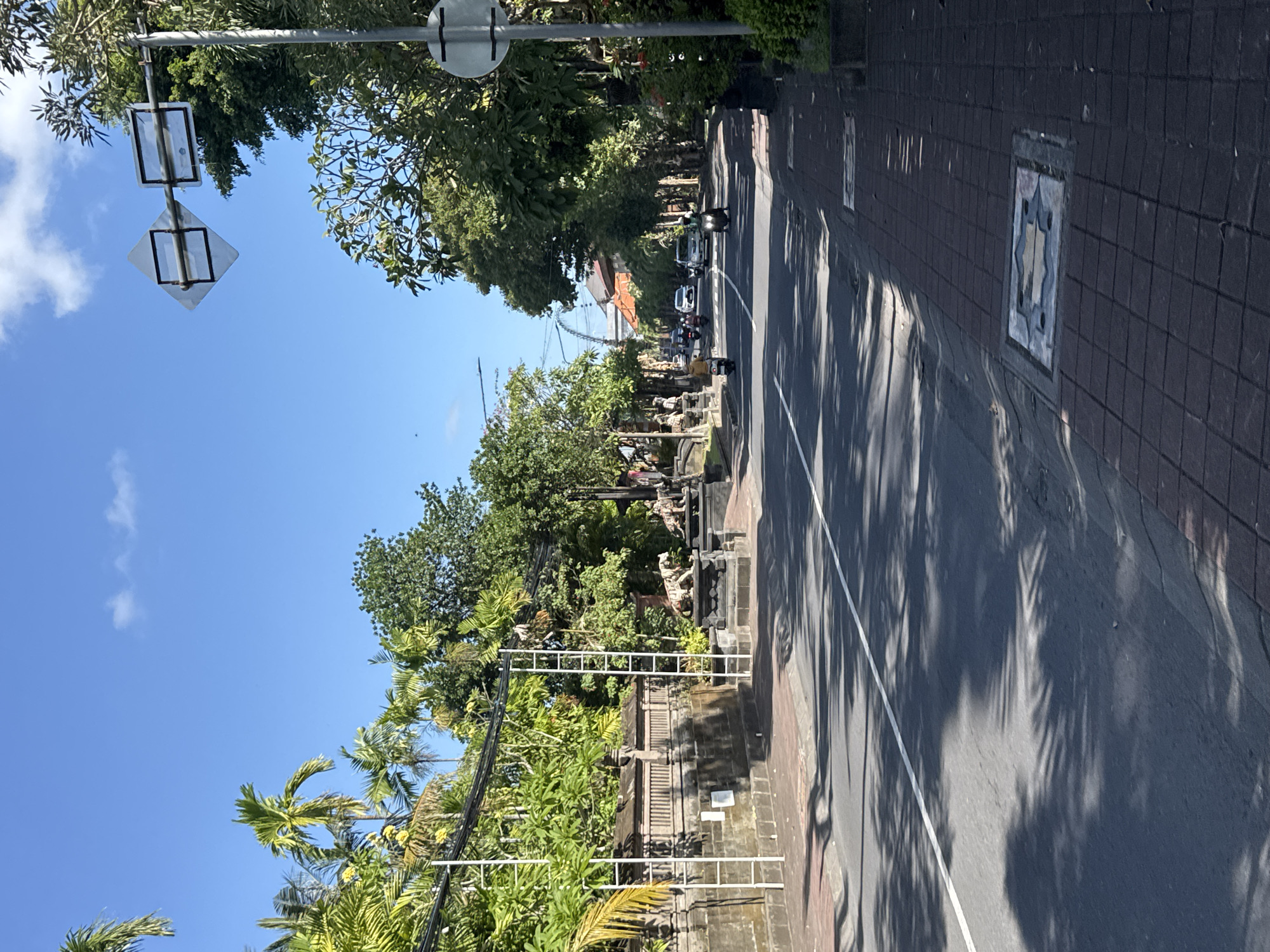In North Kuta, Indonesia, on a bright, sunny day with a clear blue sky and scattered clouds, a road curves gently to the right. The road is asphalt with a white dividing line, and palm tree shadows are cast across its surface. On the left side of the road, a stone wall topped with ornate carvings and a decorative railing runs alongside a sidewalk paved with brick. Behind the wall, lush green vegetation, including palm trees and other tropical plants, thrives. Metal scaffolding structures are erected near the wall. On the right side of the road, a brick sidewalk continues, featuring decorative square grates. A white circular sign with black horizontal lines is mounted on a pole. Further down the road, traffic can be seen, including several motorcycles and cars. A distinctive arched bridge structure is visible in the distance. The scene depicts a well-maintained urban street in a tropical environment.