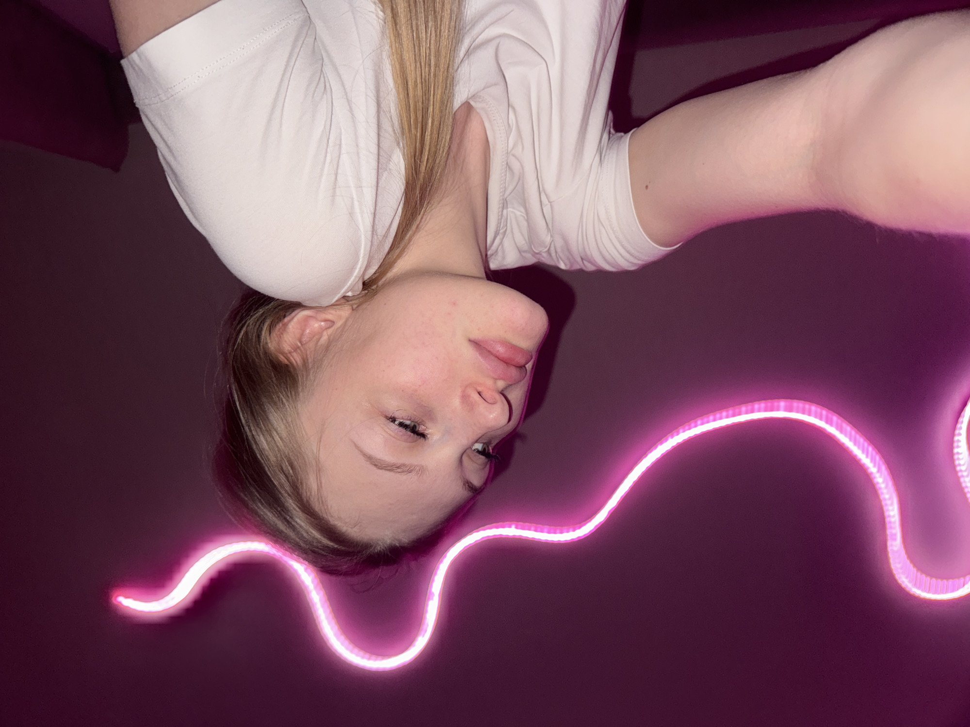 A young Caucasian female with light skin and blonde hair is captured in a selfie. She is wearing a white t-shirt. Her head is tilted upwards and to the right, with her eyes looking in the same direction. Her lips are slightly parted, and her chin is resting on her chest. A neon pink light strip snakes across the background, creating a wavy pattern against a dark purple wall. The image is cropped to focus on the upper body of the individual. The setting does not provide any identifiable features of the city of Perm, Russia.