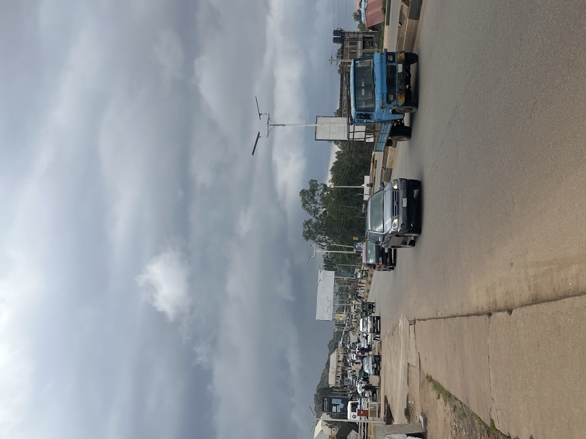 The media file depicts a busy street scene in Gura Top, Nigeria. The perspective is from a low angle, looking upwards towards the sky which is filled with heavy, dark clouds, suggesting an overcast or stormy day. The road is paved and shows some wear and tear, with a concrete sidewalk bordering one side.
Numerous vehicles are visible, indicating moderate traffic. There are several cars, including a dark-colored SUV in the foreground, and a light-blue truck further down the road. Behind these, a procession of smaller vehicles, possibly minibuses or vans, stretches into the distance. The presence of numerous vehicles suggests a bustling urban or semi-urban environment.
Buildings and structures line the street, some appearing to be shops or residential buildings, with corrugated metal roofs visible on some. There are also utility poles and streetlights, common features of an inhabited area. Trees are scattered along the roadside, some appearing to be in a lush, green state.
The scene is characterized by a sense of everyday activity, with the focus on the movement of traffic and the presence of commercial or residential structures. There are no discernible people in the immediate foreground, but the vehicles imply people are present within them. The overall mood is neutral, reflecting a typical day on a busy Nigerian street under a dramatic sky.
Some text is partially visible on a billboard or sign in the distance, but it is too blurry to be read. The time of day appears to be daytime, given the illumination, but the cloudy sky prevents precise determination. The location cues such as the architecture and vehicle types are consistent with a Nigerian setting.