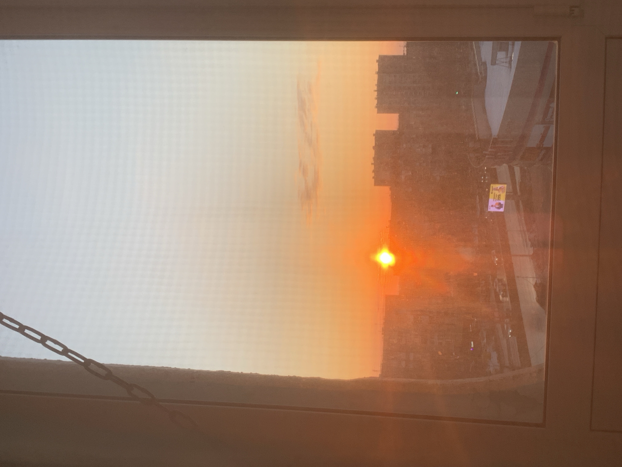 The image depicts a sunset viewed through a window, likely in an urban setting within Izhevsk, Russia. The sky transitions from a pale white to a warm orange, with the sun appearing as a bright spot on the horizon. Silhouetted against the sunset are multiple high-rise buildings, characteristic of a cityscape. Below the buildings is a road or street and some blurred signage that suggests a commercial area. The window frame is visible, with a chain hanging nearby. The view implies a high vantage point within a building.