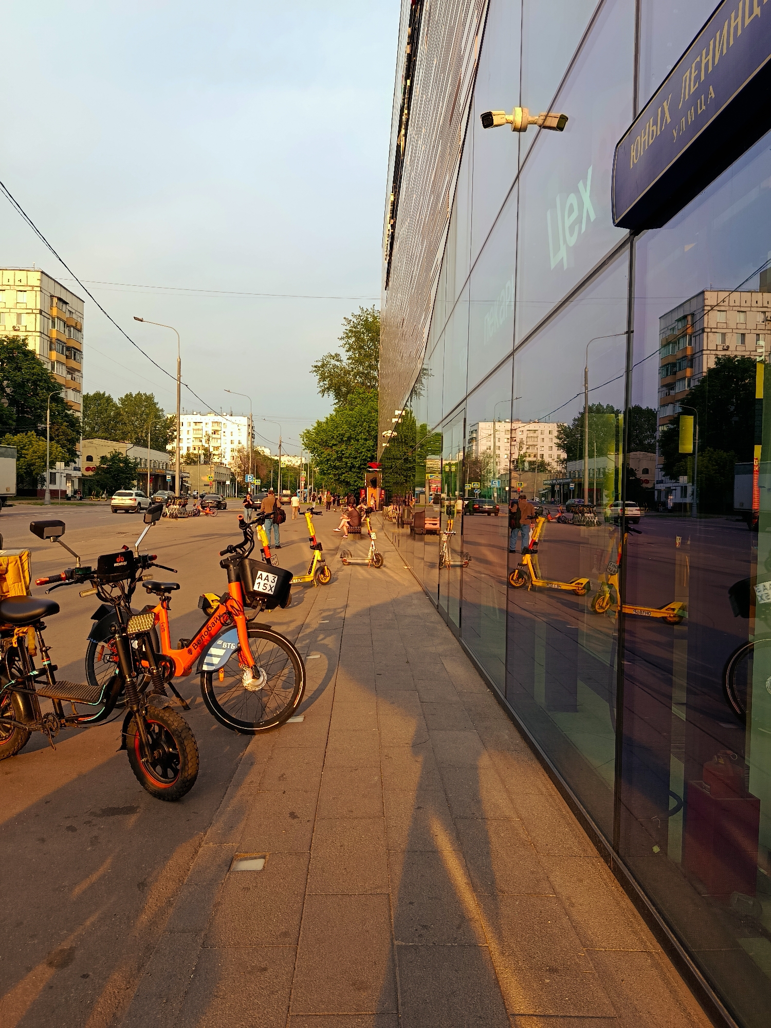 The image depicts a street scene in Moscow, Russia, with a prominent modern building featuring a glass facade on the right. The building has a blue sign with white Cyrillic lettering that reads "ЮНЫХ ЛЕНИНЦЕВ УЛИЦА" (Yunykh Lenintsev Street) and another Cyrillic word "Цех" (Tsekh). Two security cameras are mounted on the building's exterior. The sidewalk is paved with gray tiles, and several electric scooters and bicycles, many of them yellow and orange, are parked along it. In the foreground, a yellow delivery bag is attached to a black electric scooter.
Across the street, there are multi-story residential buildings and trees. The street itself is populated with cars and pedestrians. The scene is illuminated by the warm, low-angle light of what appears to be late afternoon or early evening, casting long shadows on the sidewalk. The reflections in the glass facade of the building show the street, buildings, and sky.