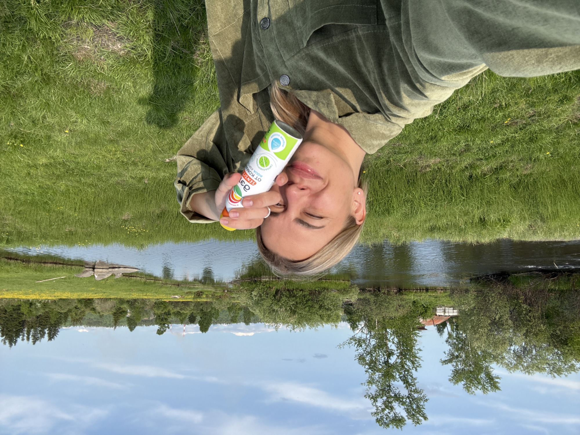 A woman is taking a selfie in a grassy area next to a body of water. She is wearing a green jacket and holding a can of spray in her right hand, which is raised to her face. She is smiling with her eyes closed. The background shows a calm river, lush green grass, trees, and a clear blue sky with scattered clouds. In the distance, there are hints of industrial structures. The location is identified as Nevon, Russia.