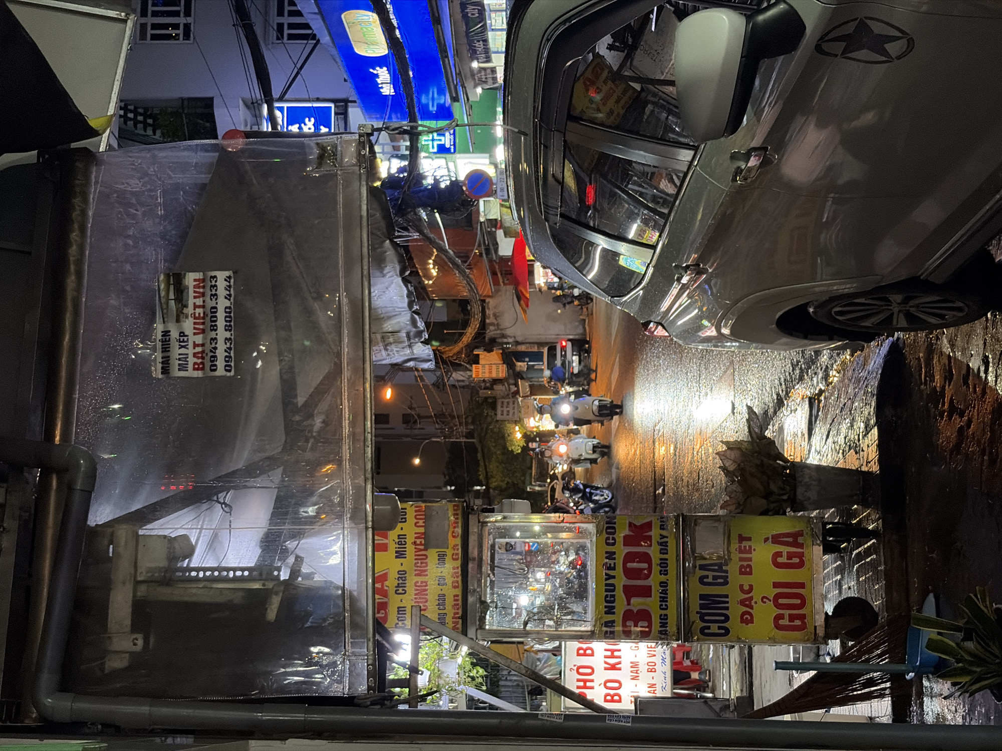 This night-time street scene, captured in Thủ Đức, Vietnam, depicts a bustling urban environment with activity centered around food stalls and traffic. The ground is visibly wet and reflective, suggesting recent rain.
In the immediate foreground, a grey car, possibly an SUV or crossover, is parked or moving slowly. A star-shaped decal is visible on its rear passenger door. The wet asphalt reflects the bright streetlights and vehicle lights.
On the left side of the street, a large, semi-permanent structure, likely a vendor stall, is prominent. It's covered with clear plastic sheeting. Several signs are affixed to it, advertising "MÁI HIÊN" (Awning) and "MÁI XẾP" (Retractable Awning), with the business name "BẠT VIỆT.VN" and two phone numbers (0943.800.333, 0943.800.444).
Further down the street on the left, several other brightly lit food stalls are visible. Their signs advertise a variety of Vietnamese dishes, including "Cơm - Cháo - Miến - Gỏi" (Rice - Porridge - Noodles - Salad), "PHỞ BÒ" (Beef Pho), "CƠM GÀ ĐẶC BIỆT" (Special Chicken Rice), and "GỎI GÀ" (Chicken Salad). One sign also lists "GÀ NGUYÊN CON" (Whole Chicken) with a price of "310K" (310,000 VND) and mentions "NHẬN ĐẶT GÀ CÚNG" (take orders for ceremonial chicken). Another sign displays operating hours from "6:00 - 22:00". A transparent display case, likely holding food items or utensils, is seen in front of one stall.
The street itself is busy with numerous motorbikes, many with riders, their headlights illuminating the wet surface. In the background, buildings with illuminated windows and blue-lit commercial signs, including a partially visible "PHARMACY" sign, line the street. A blue circular "no stopping" or "no parking" sign is also discernible. Exposed electrical wires and drainage pipes are part of the urban landscape. The scene conveys the lively atmosphere of a Vietnamese city street after dark, centered around street food and evening commutes.