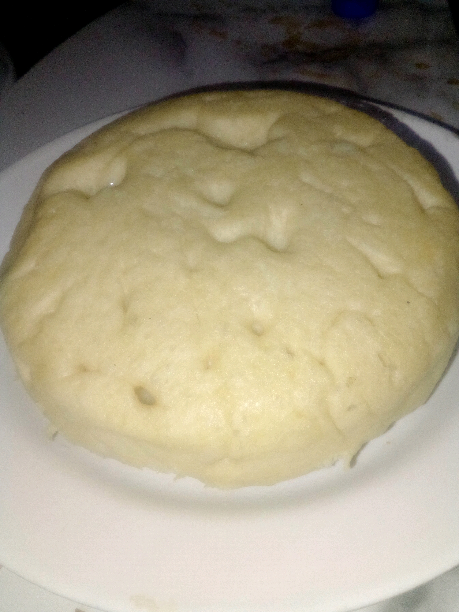 A close-up, slightly elevated shot shows a light-colored, circular food item, possibly dough or a type of bread, resting on a white plate. The food item has a slightly uneven surface with indentations and a somewhat moist sheen. The background is dark and blurred, with hints of a patterned surface and a blue object visible in the upper right corner. The overall lighting is dim, creating soft shadows and muted colors. The scene is indoors.