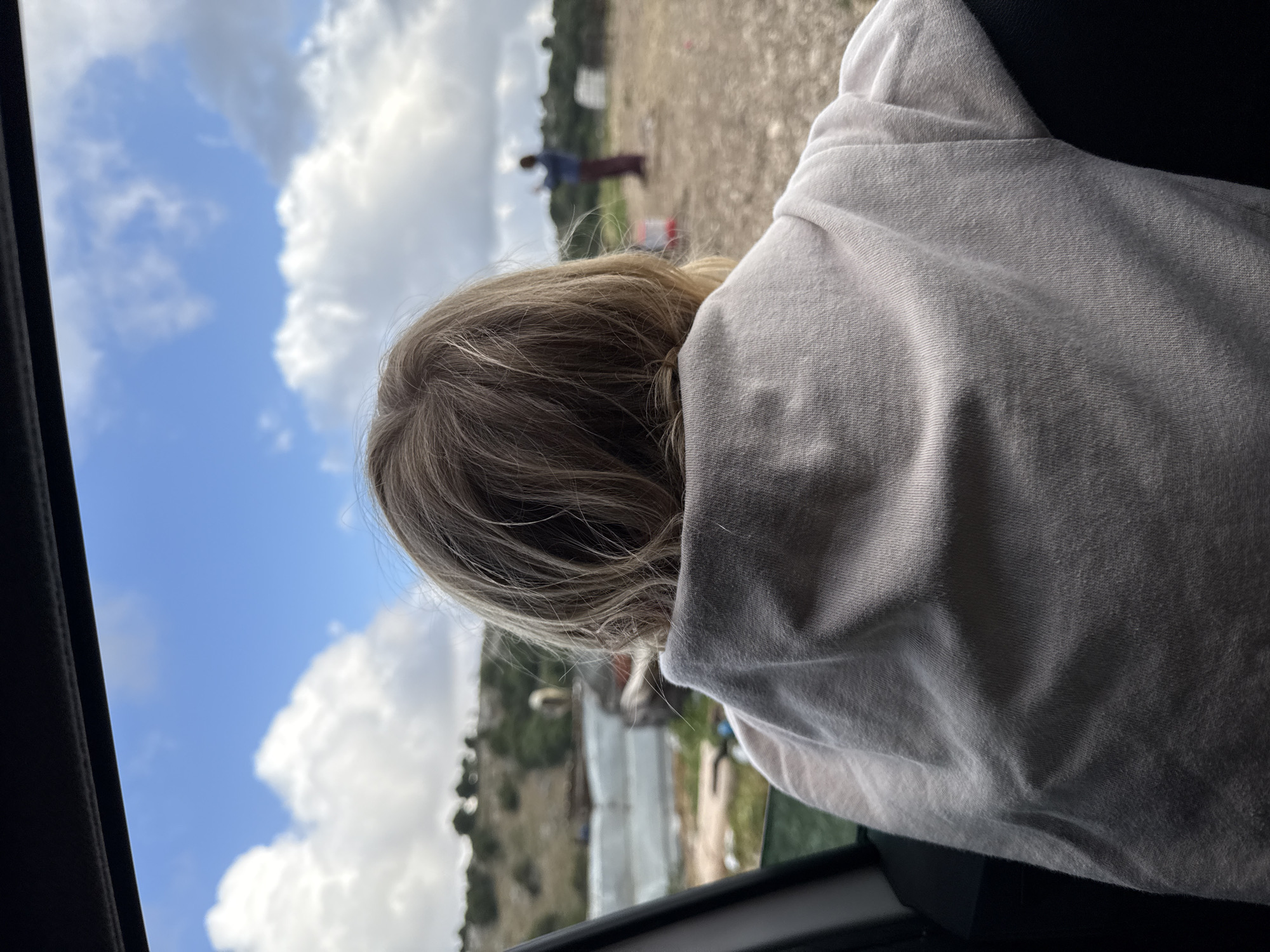 A person is looking out a car window at a landscape. The sky is blue with white clouds. In the distance, there is a figure standing on a gravelly field. There are also white structures and green vegetation. The person in the car has blonde hair and is wearing a light-colored shirt. The location appears to be Hila, Israel.