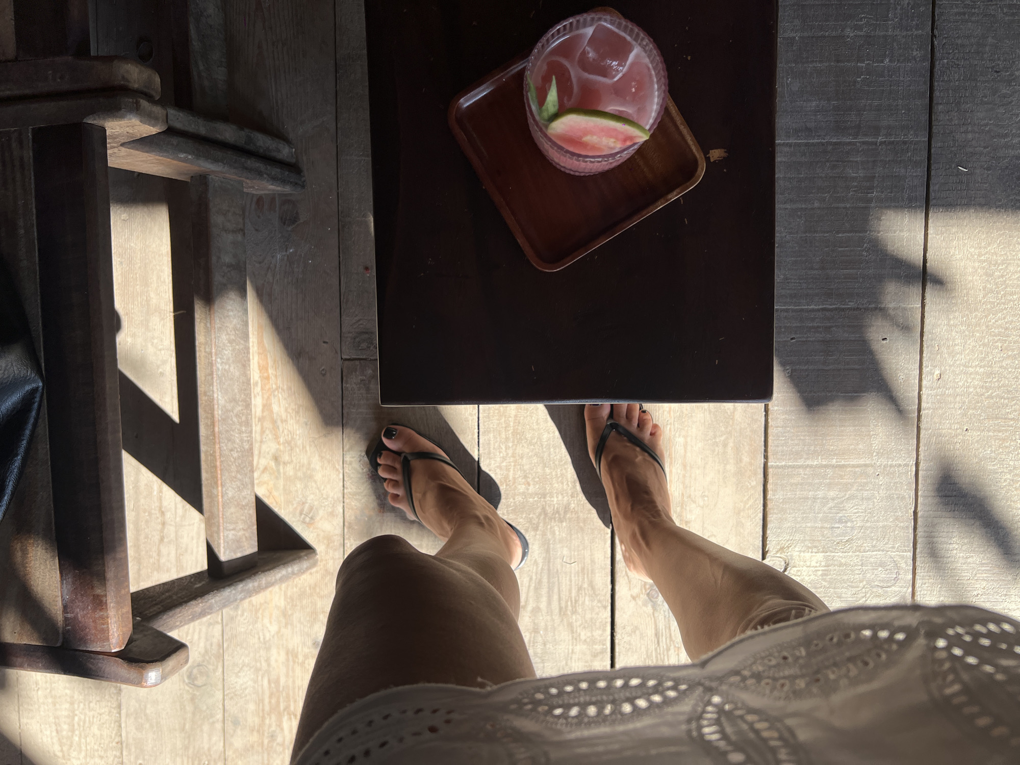 In the city of Nha Trang, Vietnam, a person is seated at a wooden table in what appears to be an outdoor cafe. Sunlight streams across the wooden floorboards, creating distinct shadows. The person, viewed from an overhead perspective, is wearing a white lace top and black flip-flops. Their legs are crossed, and their feet are visible. On the table, a dark wooden tray holds a glass of pink beverage with ice and a slice of pink fruit, possibly guava, as a garnish. The overall scene suggests a relaxed moment, possibly enjoying a drink in the late afternoon sun.