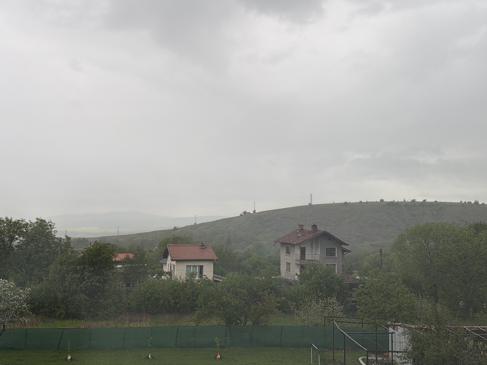 The image shows a landscape in Resilovo, Bulgaria under overcast skies. Two residential houses with red-tiled roofs are visible among greenery. A hill rises in the background, topped with a cell tower. A green fence borders a lawn in the foreground. The overall environment suggests a rural or suburban setting, typical of Resilovo's location in western Bulgaria.