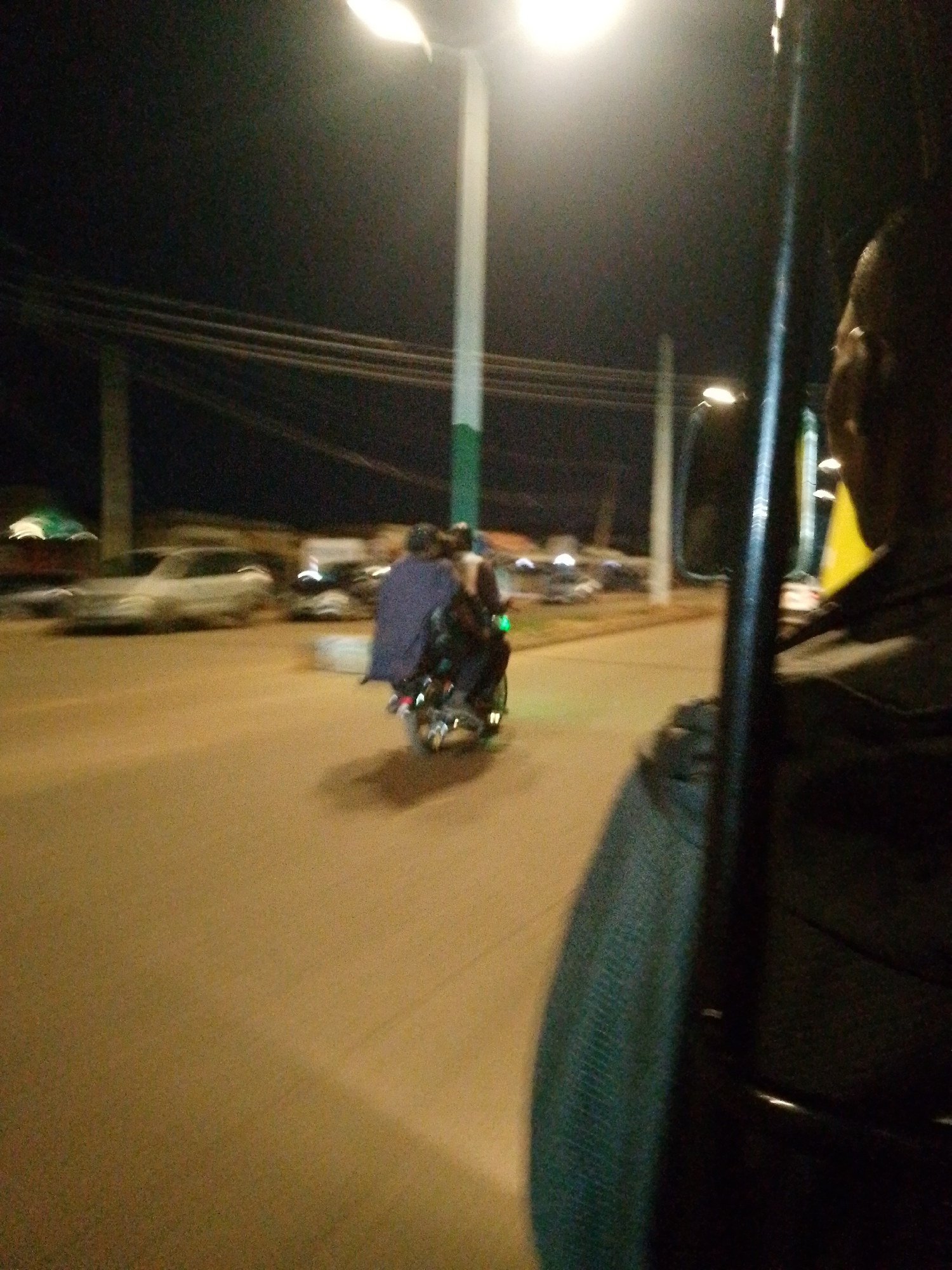 The image shows a night scene in what appears to be a street in Bukuru, Nigeria, based on the presence of streetlights and the general environment. The photo is likely taken from inside a vehicle, possibly a taxi or another mode of public transportation, as suggested by the partial view of a person in the foreground on the right side of the image. This person is wearing a dark-colored shirt or jacket. The street is paved, and there are several vehicles parked along the roadside, including cars and a motorcycle with multiple occupants. The motorcycle is moving and has a blue cloth or blanket draped over the passengers. There are overhead power lines visible against the night sky. The streetlights are emitting a bright light, illuminating the road and surrounding areas.