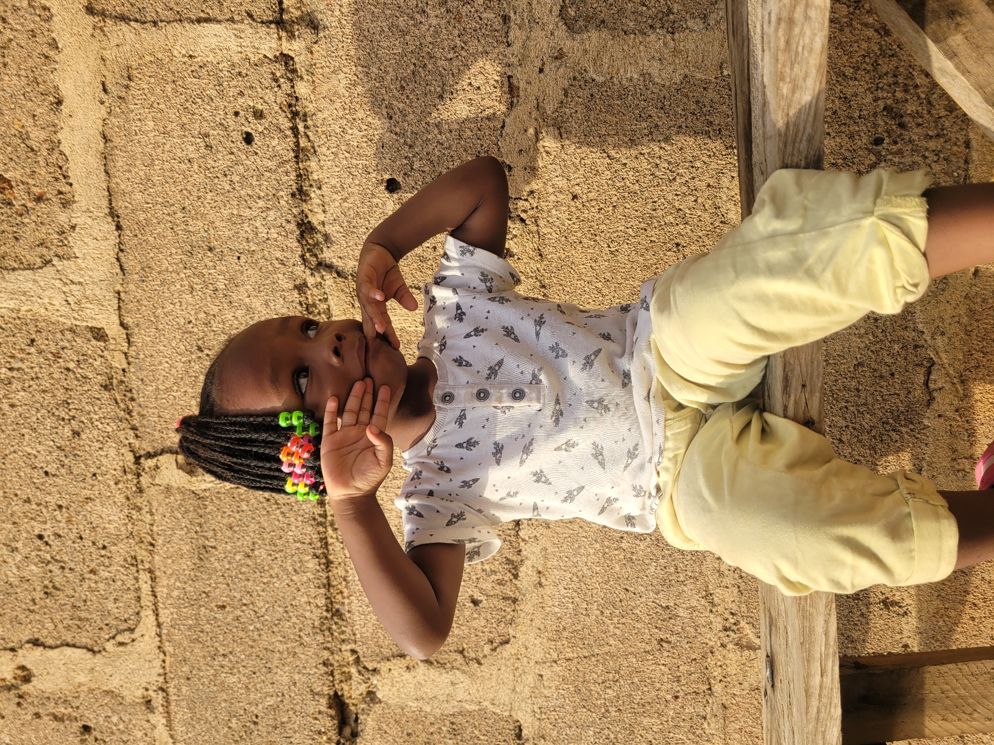 A young Black girl, wearing a white t-shirt with a repeating rocket pattern and light yellow pants, sits on a wooden bench. Her dark braided hair is adorned with colorful beads in green, orange, and pink. She is looking to her right with her hands raised to her face, covering her mouth and cheeks. The background consists of a rough, textured, light brown stucco wall. The scene appears to be outdoors, likely in the city of Barakin Akawo, Nigeria, given the architectural style of the wall and the natural lighting suggesting a warm climate.