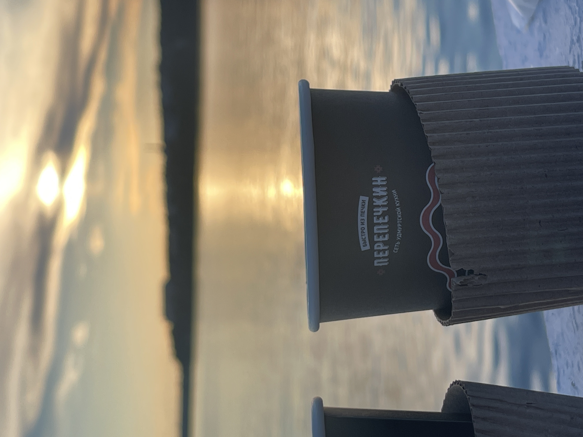 The image shows two disposable coffee cups with cardboard sleeves in the foreground, positioned on a white surface. The cups are dark gray with white lettering and a red wavy line design. The lettering on the cup in the foreground reads "БЫСТРО ИЗ ПЕЧКИ" and "ПЕРЕПЕЧКИН" with "СЕТЬ УДМУРТСКОЙ КУХНИ" below it. In the background, a body of water reflects the golden light of a sunset or sunrise. The horizon is lined with a dark silhouette of land, possibly a city skyline or treeline. The sky is filled with clouds, with the sun shining brightly through them. The overall scene evokes a calm and serene atmosphere, likely taken outdoors by a waterfront at dusk or dawn.