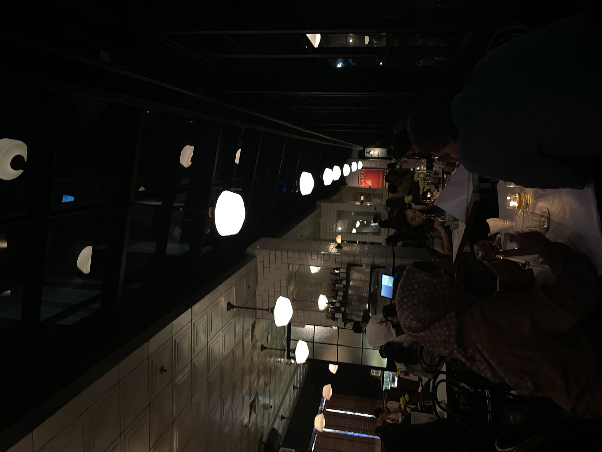 The image depicts the interior of a restaurant, possibly located in Central Jakarta, Indonesia, based on visual cues such as architectural elements and potentially the cultural context. The lighting is dim, primarily provided by numerous hanging spherical lights that cast a warm glow. The walls are covered with white, rectangular tiles arranged in a pattern, possibly a grid or staggered brick style. People are seated at tables covered with white tablecloths, suggesting a formal or upscale dining experience. Some diners are visible, and they seem to be engaged in conversation and enjoying their meals. Table settings include glassware, candles, and possibly floral arrangements. A bar area with shelves displaying bottles is visible in the background.