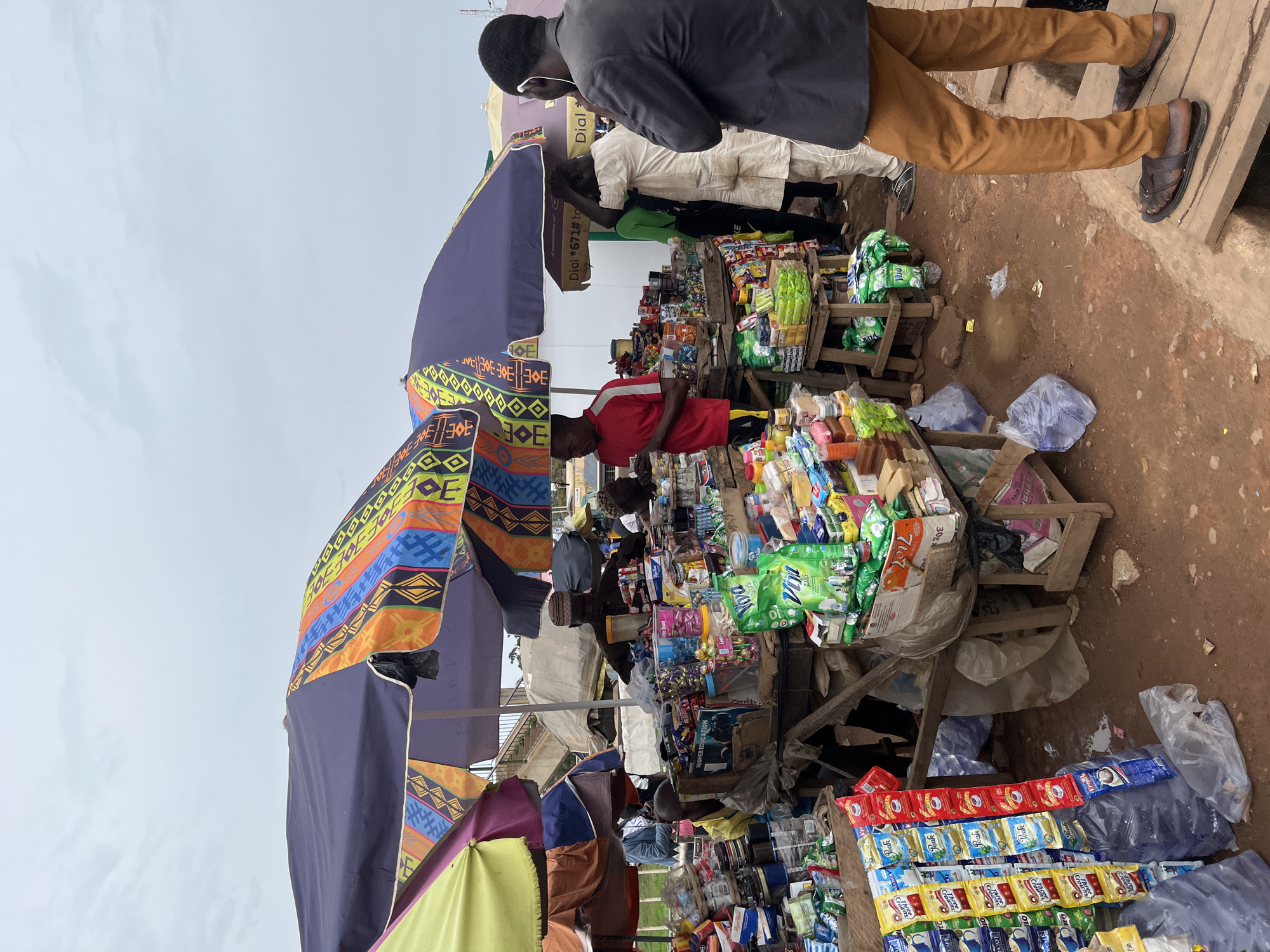 The scene is a bustling marketplace in Gura Top, Nigeria. Stands and small tables are laden with merchandise, including packets of food items, soaps, and possibly other household goods, all meticulously displayed. Several people are visible, including a man in a red shirt who appears to be tending to his display of goods. Other individuals are seen observing the merchandise. The environment is characterized by a mix of open-air stalls shaded by umbrellas, adding to the vibrancy of the market setting. The ground appears to be an earthen surface, indicative of an informal or semi-permanent trading area.