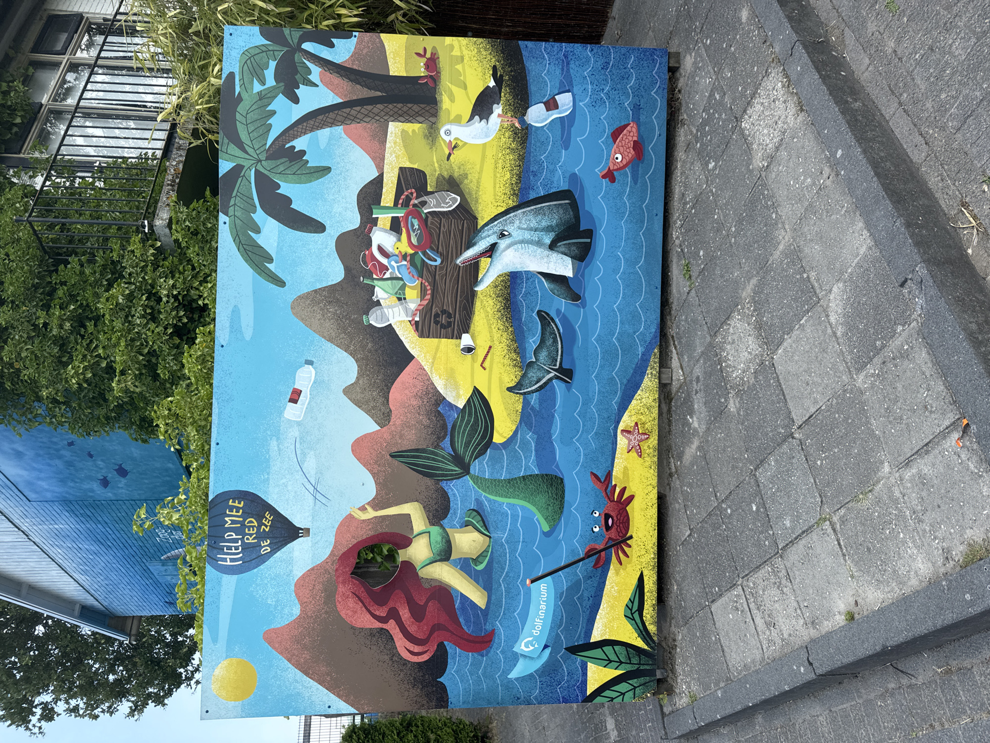 The image shows a colorful mural displayed outdoors, mounted on a wooden structure against a paved walkway. The mural depicts a marine-themed scene with a mermaid cut-out, marine life, and a message about saving the sea. The background features stylized mountains, palm trees, and a sunny sky. In the foreground, there's a representation of a beach with sand, plants, a crab, and a starfish. The sea area of the mural shows a dolphin, a seagull, a whale tail, and various plastic waste items like bottles and bags, all within a blue water environment. A banner on the mural reads "dolfinarium". The mural is positioned on a sidewalk in front of a building with a balcony and trees. The text on the mural also includes "HELP MEE RED DE ZEE," which translates to "HELP ME SAVE THE SEA." This mural is located in the city of Harderwijk, Netherlands, specifically at the Dolfinarium Harderwijk, an animal park focused on marine mammals.