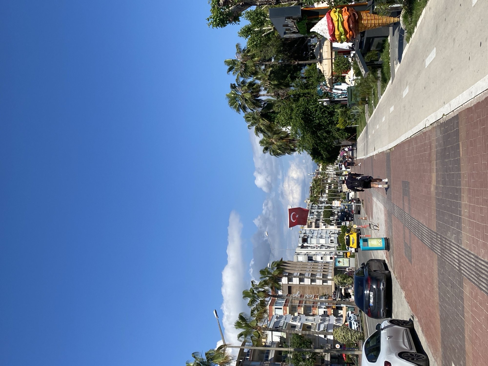 The image depicts a street scene in Alanya, Turkey. The sky is a clear blue with a few scattered clouds. A Turkish flag is prominently displayed. Buildings line the street with palm trees interspersed. A paved walkway runs alongside the street, with a bicycle lane visible. A pedestrian is walking along the walkway. Several cars are parked along the street. A large ice cream cone sculpture stands near a building.