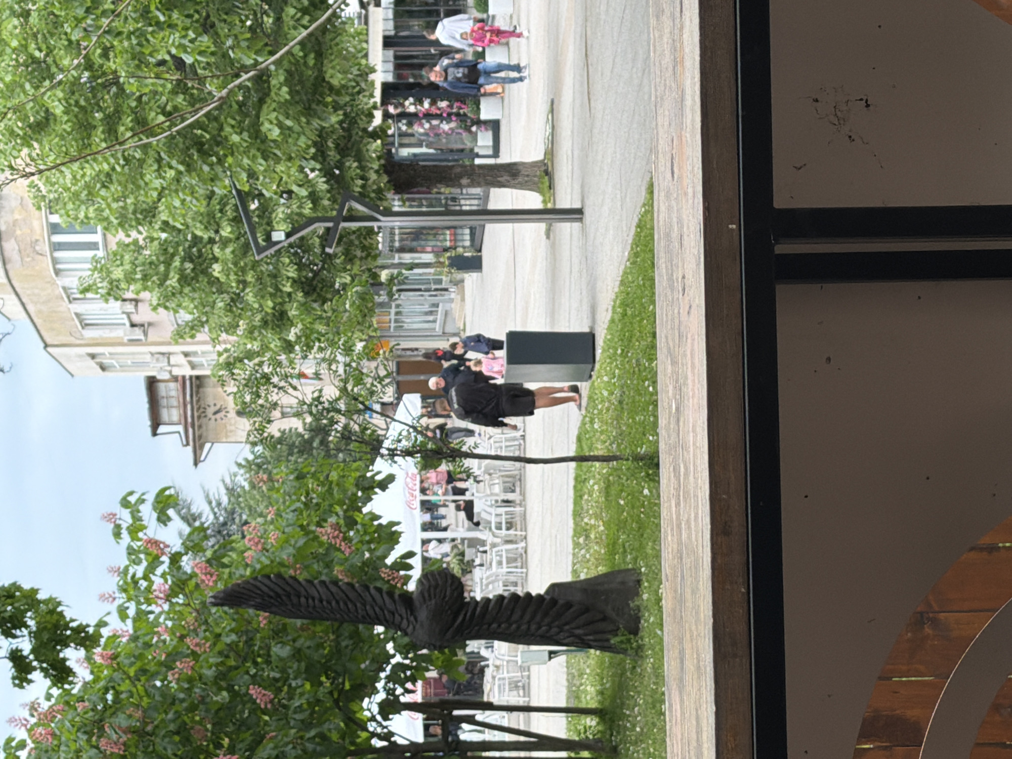 The image depicts an outdoor scene in Vratsa, Bulgaria. In the foreground is a dark wooden sculpture of an eagle, positioned amongst flowering trees. Behind the sculpture is a grassy area where a person in dark clothing stands using their cell phone. Further back is an outdoor café area with white chairs and tables, situated under Coca-Cola branded awnings. In the background, people are seen walking on the sidewalk, and a building with a light-colored façade can be observed. The image is taken from an angle, possibly from inside a building or structure with a wooden frame.