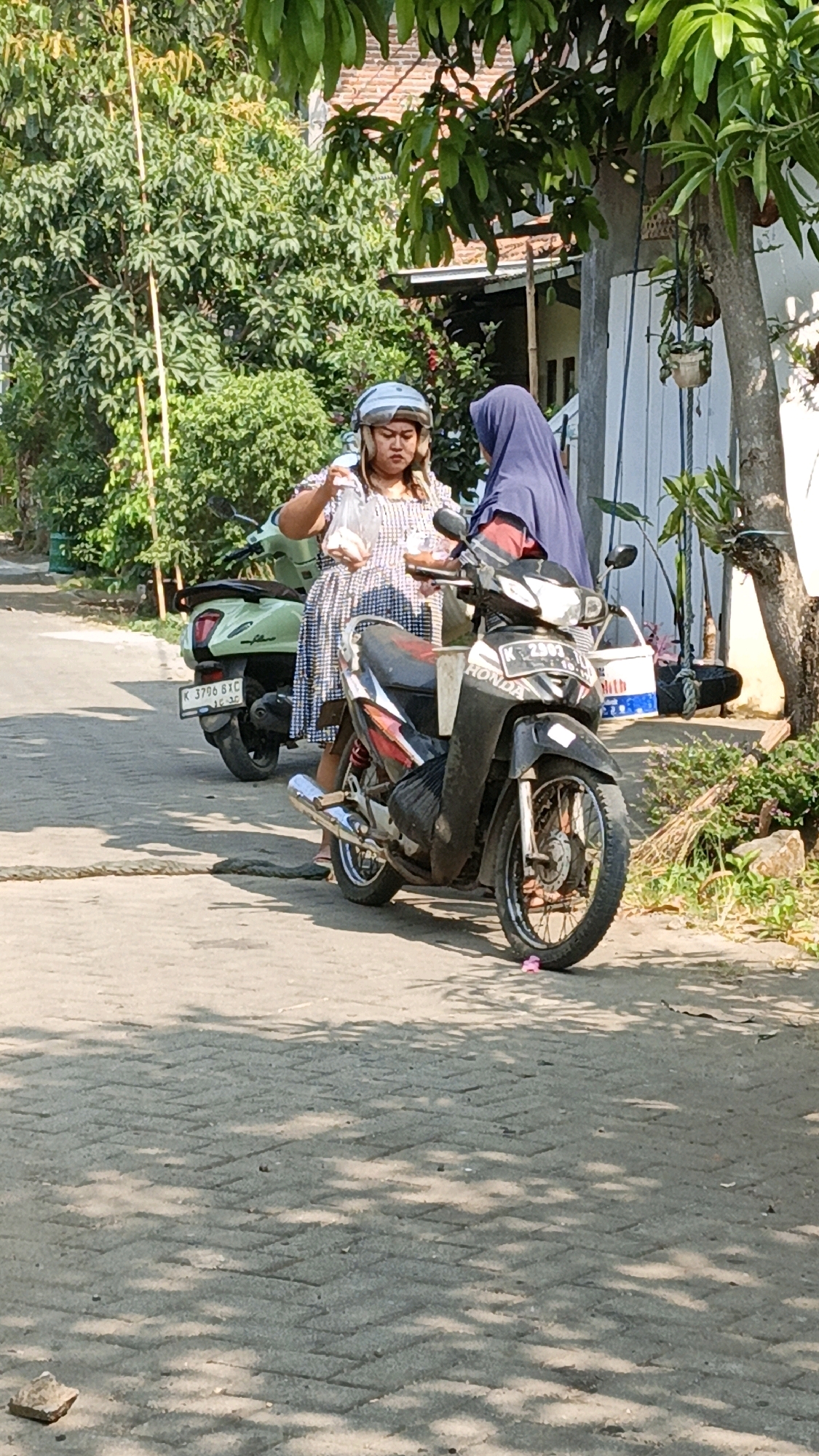 Two women stand next to a black Honda motorcycle parked on a paved street in Cikal, Indonesia. The woman on the left, wearing a checkered dress and a light blue helmet, holds a plastic bag with items inside. The woman on the right wears a dark blue hijab and a red top. A light green scooter is parked behind the motorcycle. The environment is leafy and appears residential, with a building visible in the background. A rope lies across the street in the foreground.