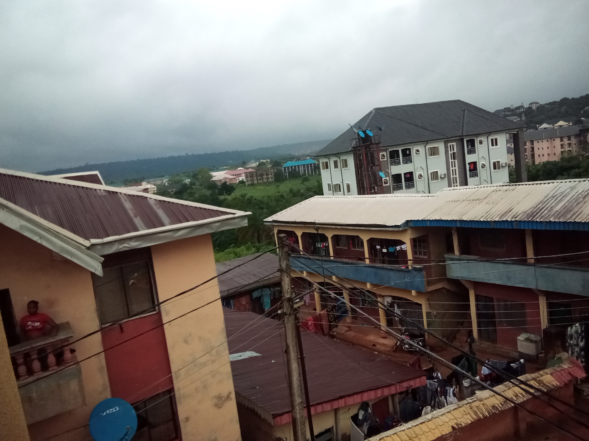 The image depicts a densely populated urban area in Amaokpala, Nigeria, characterized by multiple residential buildings and an overcast sky. In the foreground, a two-story building with a reddish-brown corrugated metal roof is visible on the left. A person in a red shirt is standing on a balcony of this building, leaning on a railing. A blue satellite dish is mounted on the exterior wall below the balcony.
Adjacent to this building is another structure with a similar reddish-brown metal roof and a yellow facade on its lower level. Above this, a multi-story building features a blue trim along its upper balcony and a light yellow facade. Laundry is hung out to dry on balconies and clotheslines across several buildings, indicating residential activity. Wires from a utility pole are prominently strung across the scene, connecting various structures.
Further in the background, a larger, modern multi-story building with a dark gray roof and light-colored walls stands out. This building has multiple windows and balconies, with some laundry visible. In the distance, more buildings are scattered across a green, hilly landscape, with a hazy, mountainous horizon under a cloudy sky. The overall impression is one of a lived-in, working-class urban environment with signs of daily life unfolding amidst the residential structures.