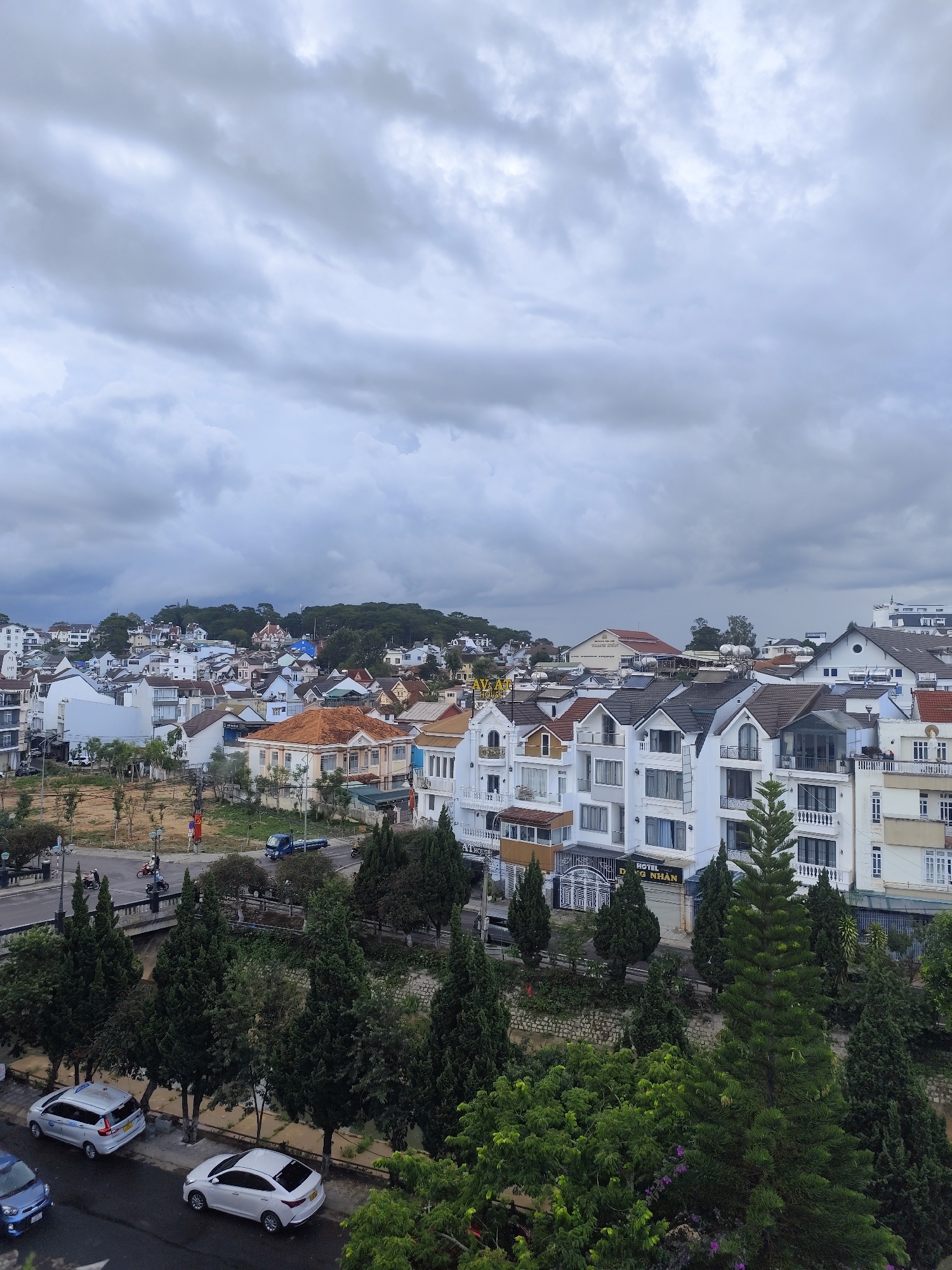 The image depicts a cityscape in Da Lat, Vietnam, under an overcast sky. The foreground features dense greenery with various trees. A street runs horizontally, with several parked cars and a few motorcycles. The middle ground is dominated by a cluster of buildings, predominantly white with brown and grey rooftops. The buildings appear to be a mix of residential and commercial properties, including "HOTEL G NHAN" as visible on one building. In the background, more buildings are seen on a distant hillside covered with trees. The overall scene suggests a residential area within the city.