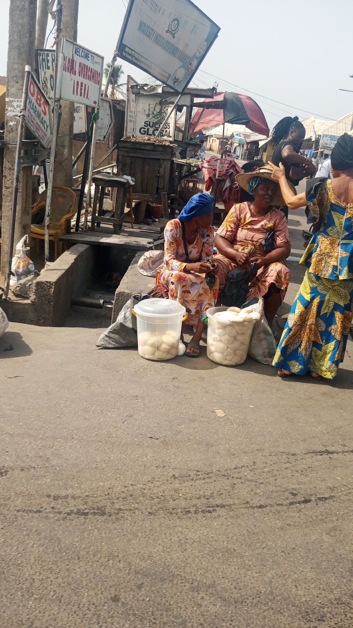 The image depicts a bustling street market scene in Lokoja, Nigeria, under bright daylight with a clear sky. In the central foreground, two women are seated on a low concrete ledge beside a paved road. One woman, wearing a blue headscarf and a multicolored patterned dress, is looking down at her hands. Next to her, another woman in a wide-brimmed straw hat and a patterned dress looks towards the right. In front of them are two large, clear plastic buckets filled with numerous light-colored, rounded food items, likely bread rolls, suggesting they are vendors.
To the right, a third woman, partially visible, stands in a vibrant blue and yellow patterned dress, seemingly interacting with the woman in the straw hat. Further back, another woman with dark hair is visible near some market stalls.
The background reveals a typical market environment with makeshift stalls, wooden structures, and various goods. Several signposts are prominently displayed. Readable text includes: "OBS PHARMACY," "WELCOME TO GLOBAL OVERCOMERS ARENA," and "MAJESTY INTERNATIONAL COLLEGE" with its logo. Another sign near a wooden stall reads "SUCCESS GLOBAL." The overall setting is a vibrant, active marketplace, characterized by vendors, customers, and a mix of permanent and temporary structures. The ground is a mix of paved road and rough concrete.
