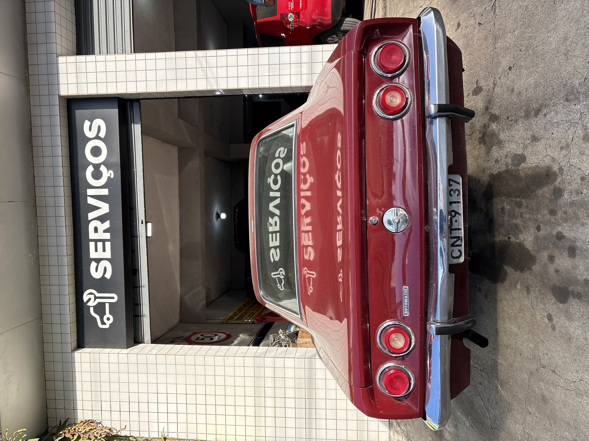 This is a rear-view, medium shot of a vintage, dark red coupe parked outside a garage with a tiled exterior. Above the garage entrance, a black sign with white lettering reads "SERVIÇOS" accompanied by a white icon of a car and a wrench, indicating it's a service or repair shop. The car has four circular taillights, two on each side, and a chrome bumper. The license plate is visible, displaying "CNT-9137". Inside the garage, another red vehicle, possibly an SUV, is partially visible. The ground is concrete, with some oil stains. The environment appears to be a sunny day, with shadows cast on the ground. There are no people clearly visible in the image. The presence of the license plate from São Paulo, Brazil, as indicated by the CNT prefix, suggests the location of this scene.