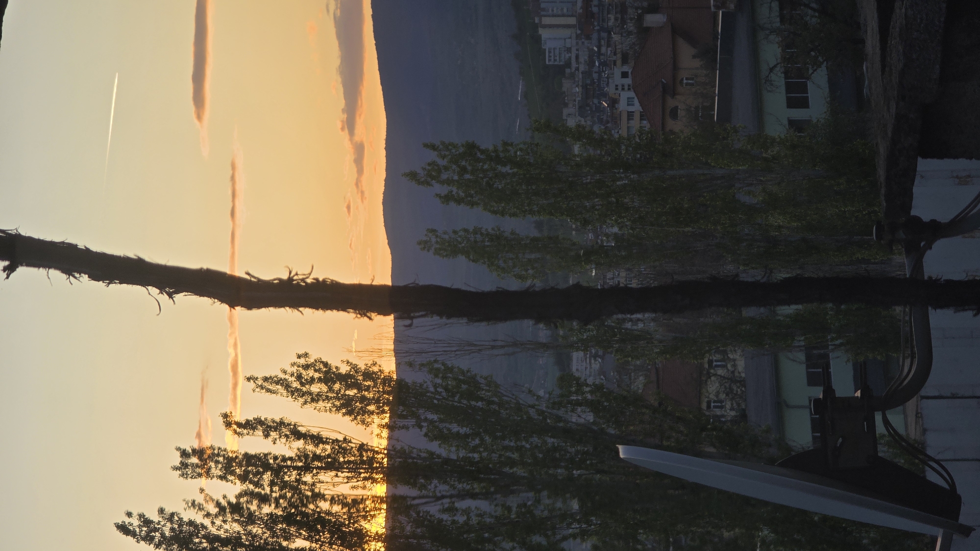 The image shows a sunset over Blagoevgrad, Bulgaria. A satellite dish and some trees are in the foreground. In the background, a mountain range is visible, with the city nestled in the valley. Contrails from a plane streak across the sky. The scene is reflected, indicating the image may have been rotated.