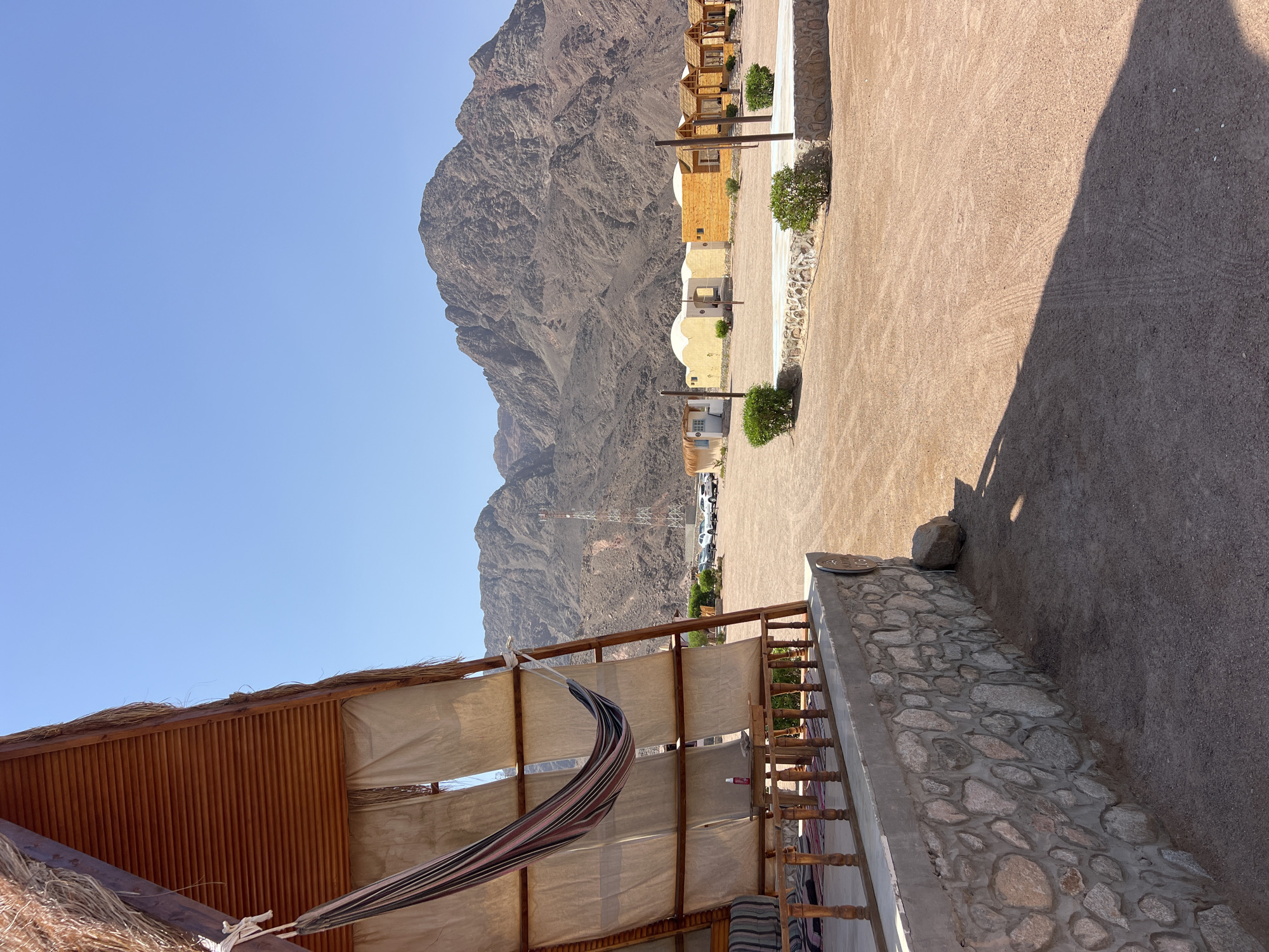 The image depicts a sun-drenched, arid landscape with a clear blue sky. In the foreground, a triangular-shaped structure with a thatched roof and wooden beams is visible on the left. A striped hammock hangs from the beams of this structure, and a canvas curtain drapes down. A low stone wall with concrete mortar supports the front of this structure. A small table with a bottle of spray sits on a wooden railing.
Beyond this structure, the ground is covered in loose, reddish-brown gravel. Several low-lying buildings with light-colored, dome-shaped roofs and a few wooden structures with thatched roofs are arranged in a semi-circle. A dirt road or pathway is visible, with tire tracks imprinted on the surface. A telecommunications tower stands in the middle ground. In the background, a large, rugged, rocky mountain range dominates the horizon. Several parked vehicles, including cars and what appears to be a utility vehicle, are visible in the distance. The overall scene suggests a remote desert resort or camp. The specific location is described as Unknown, Egypt. There are no people visible in the image.