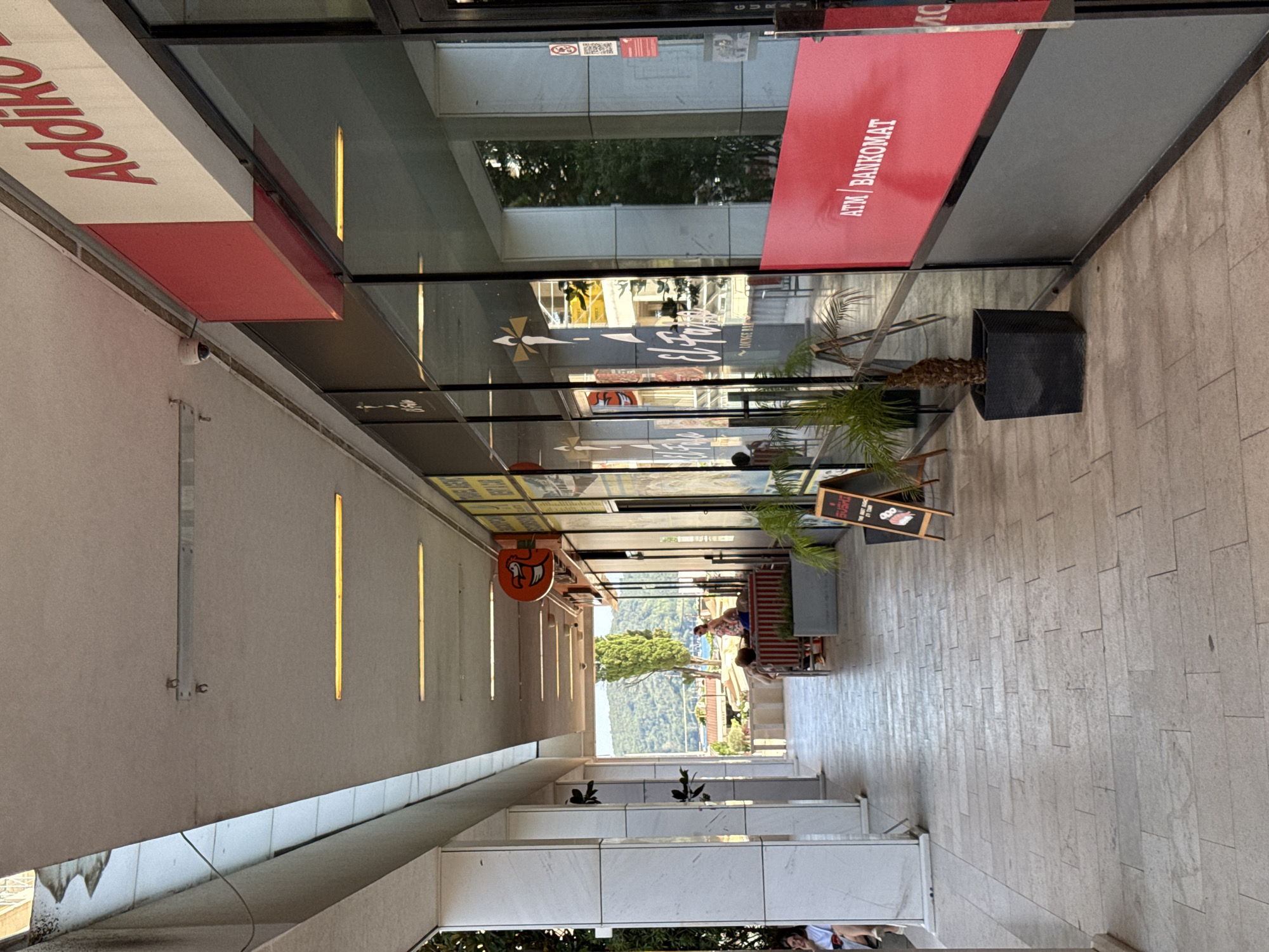 The image, taken in Budva, Montenegro, depicts a covered walkway with a tiled floor. The walkway is lined with storefronts and windows. On the left side, a sign with red lettering reads "Addiko Bank." To the right, a red awning displays white text that says "ATM BANKOMAT." Further down the walkway, a person is seated on a bench with plants nearby. In the background, a view of trees and hills suggests an outdoor setting. The lighting indicates it is daytime. The scene appears calm and relaxed, with no overt signs of activity. Reflections in the windows offer glimpses of the environment and other people.