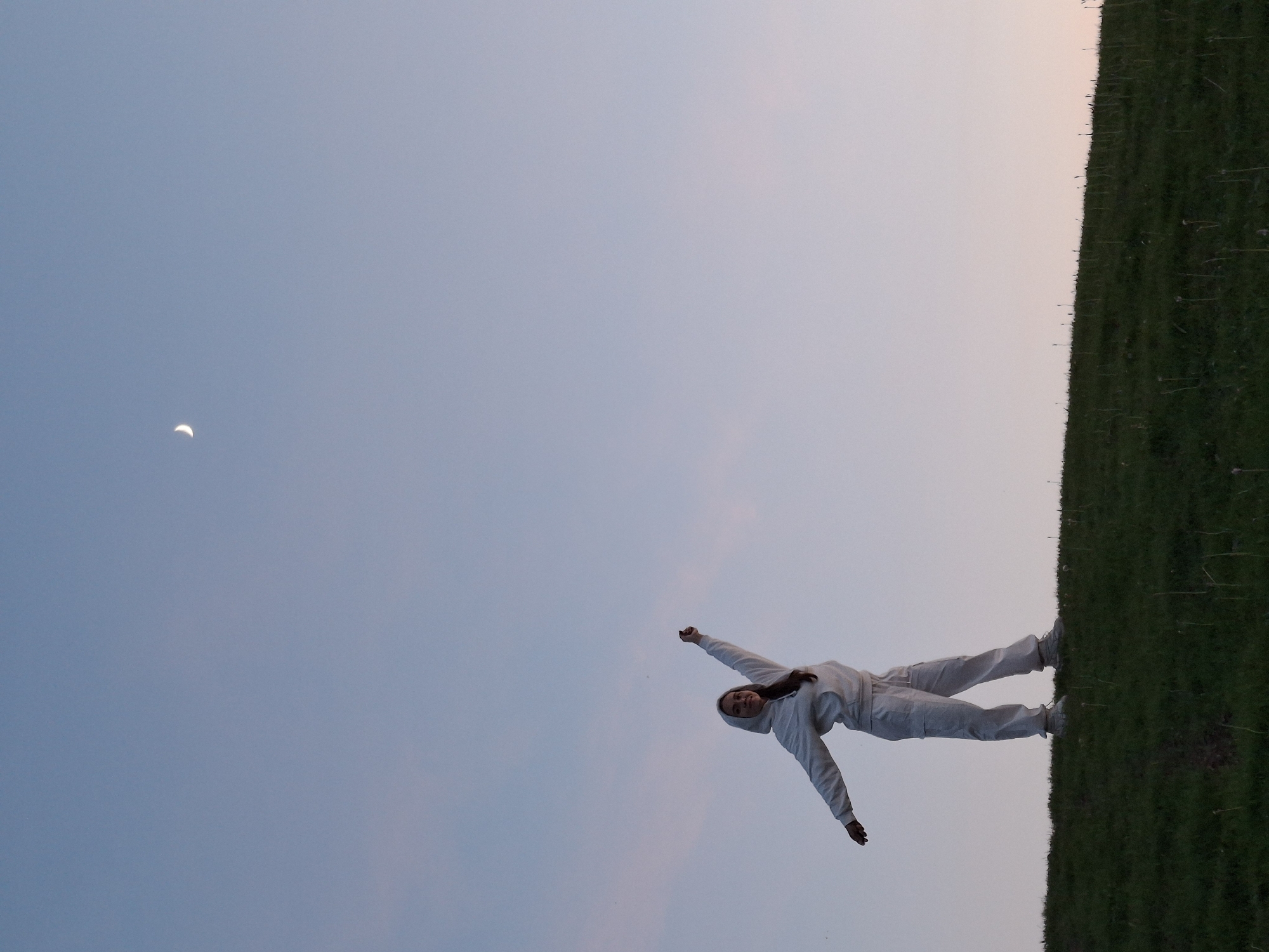 A photograph shows a person in light-colored clothing lying on a grassy slope under a twilight sky with a crescent moon. The location could be in Ольховка, Russia, given the request for city context. The individual is prone with arms outstretched. The scene suggests a quiet outdoor setting during twilight.