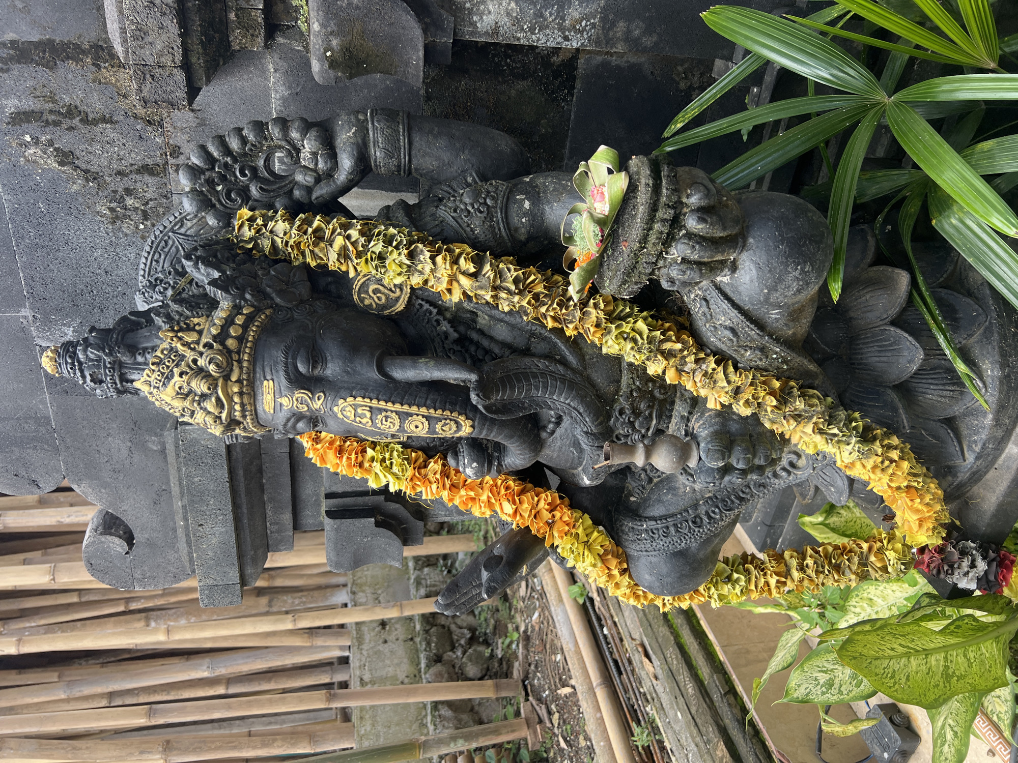 The image shows a stone statue of a deity, likely Ganesha, adorned with garlands of yellow and orange flowers. The statue is situated outdoors near lush green plants, with a bamboo fence visible in the foreground. The location appears to be in the Ubud District of Indonesia, based on the style of the statue and the surrounding environment.