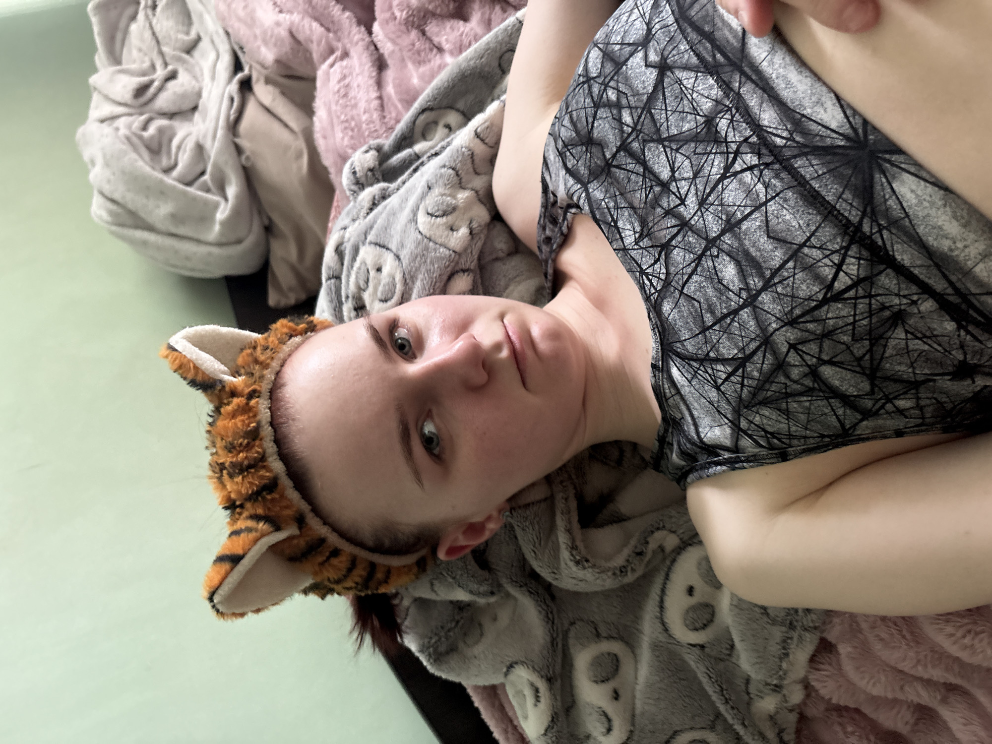 A young woman with light skin and blue eyes wears a headband with tiger ears. She has dark hair pulled back. She is lying down on a bed covered with blankets, one grey with a panda pattern and another pink and fuzzy. Behind her, a light green wall is visible. She is wearing a grey tank top with a black geometric pattern. Her expression is neutral.