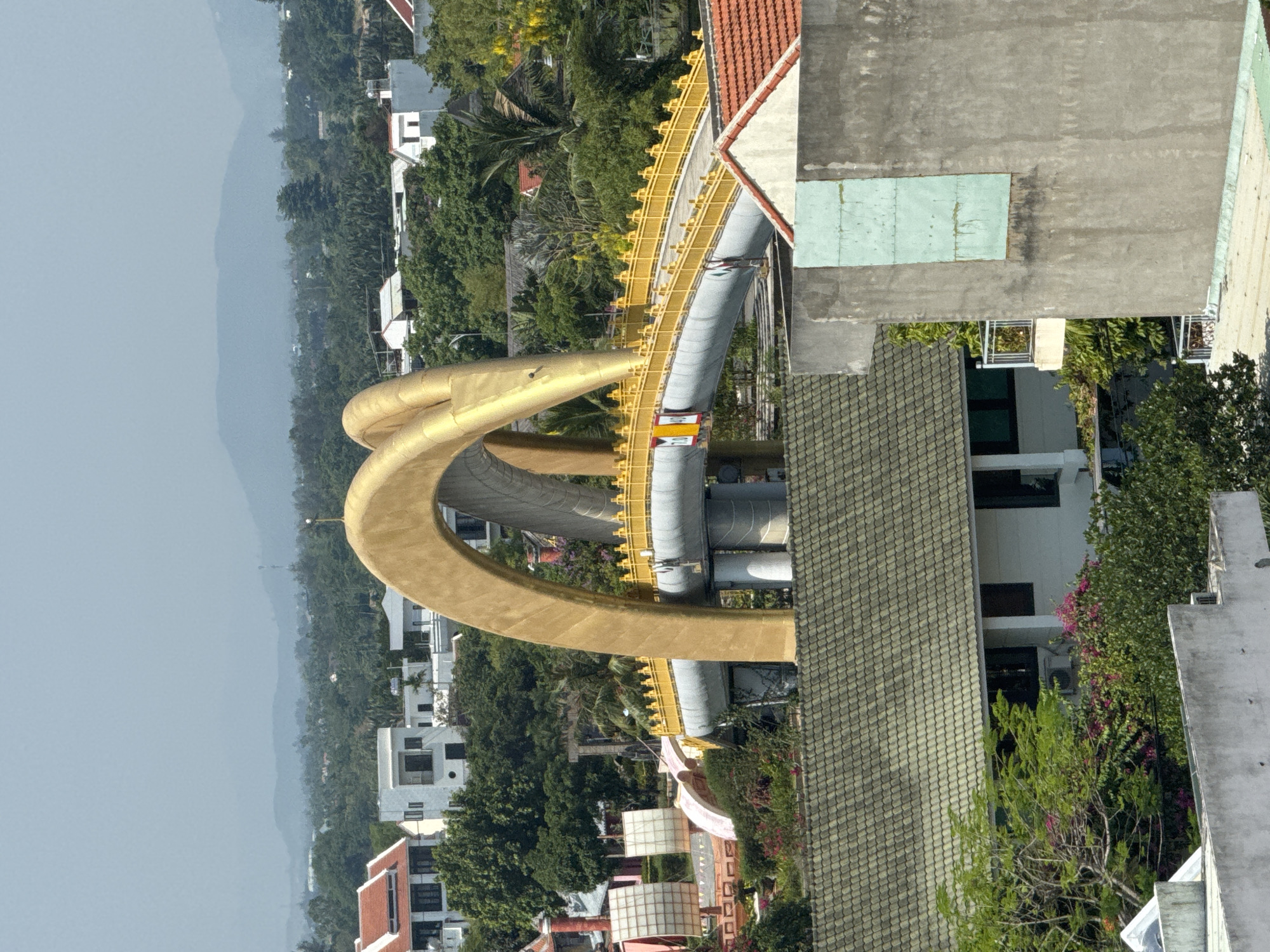 The image shows the Cau Tran Thi Ly Bridge in Đà Nẵng, Vietnam. The bridge has a distinctive loop shape and is painted gold. It is surrounded by buildings, trees, and a distant mountain range. The photo appears to have been taken from a high vantage point, potentially from a nearby building. The sky is overcast. The bridge carries gray pipeline structures. The foreground contains rooftops of adjacent buildings. There are no people visible in the image.