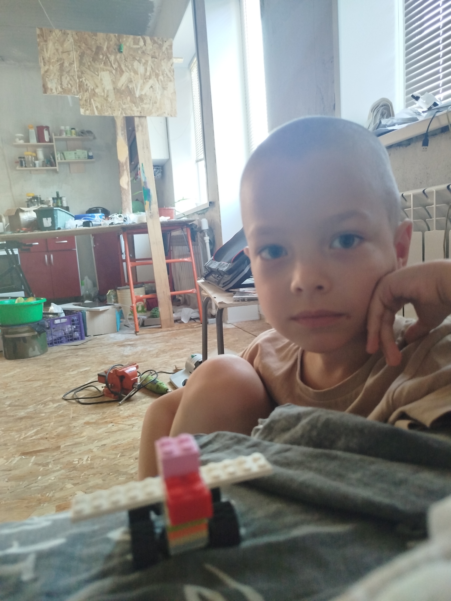 The image depicts a close-up of a young boy's face, looking directly at the camera with his left hand resting on his cheek. He has a shaved head and is wearing a light brown t-shirt. In the foreground, a partially constructed toy made of Lego bricks is visible. The background appears to be an indoor setting, possibly a workshop or a room under renovation, with wooden flooring and walls, various tools, shelves with containers, and a large window with blinds. The overall impression is that of a casual, possibly domestic, environment. The location is not ascertainable from the image itself, and therefore no details about Zavodoukovsk, Russia can be provided.