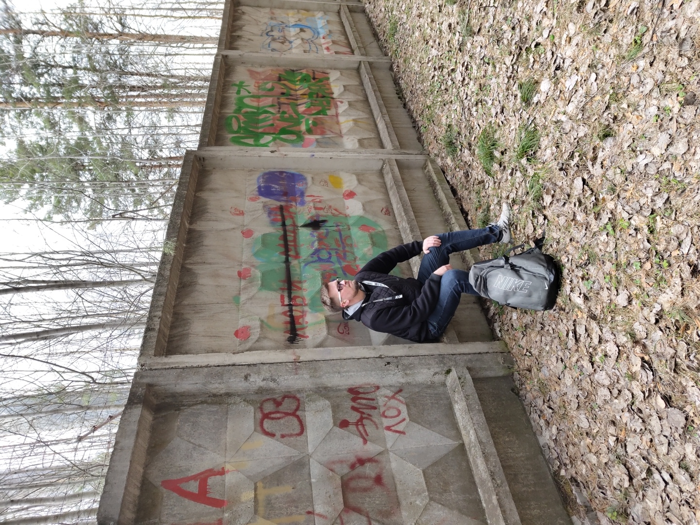 The image depicts a man sitting on the ground next to a tall, graffiti-covered concrete wall in an outdoor setting, likely in Прикамский, Russia. The man wears a black hooded jacket, blue jeans, and light-colored sneakers, with sunglasses resting on his head. He is in a relaxed posture, with his legs bent. A grey Nike duffel bag, clearly displaying the white "NIKE" logo, rests on the ground beside him.
The ground is covered with a thick layer of dried, fallen leaves and sparse patches of grass, indicating a season such as late autumn or early spring. The concrete wall, constructed from large, rectangular panels, some with a textured or faceted pattern, is heavily adorned with a variety of colorful graffiti. Visible text and symbols include red "A" and possibly Cyrillic characters, alongside abstract designs and splatters in green, blue, yellow, and black.
Behind the wall, numerous bare-branched deciduous trees and some evergreen pines extend upwards, suggesting a forested or treed area. The sky above is bright but overcast. The scene is illuminated by daylight.