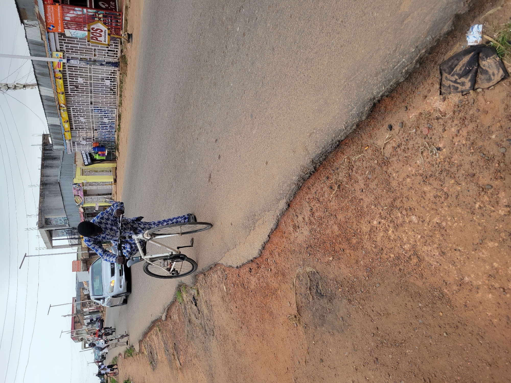 The image shows a street scene in what appears to be a built-up area. A person is riding a white bicycle on the road. There is a silver car partially visible, heading in the same direction as the cyclist. Other vehicles, including motorcycles, are further down the road. Buildings with shops and commercial establishments line the street. A "POS Available" sign is visible on one of the buildings. The road surface is paved, but the edges are rough and the shoulder appears to be dirt. There's a piece of black cloth or debris on the shoulder. The sky is overcast. The location is the city of Unknown, Unknown.