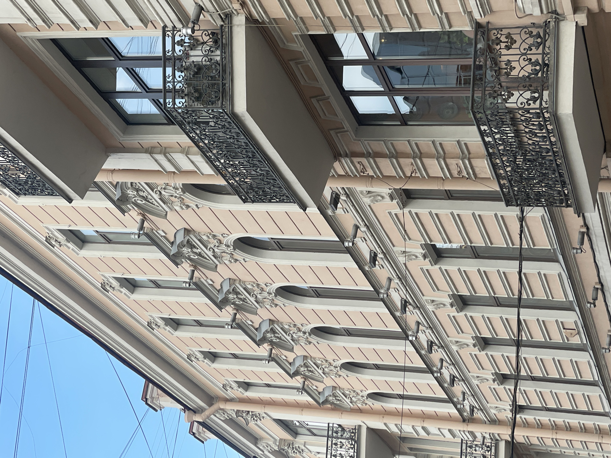The image shows a building facade in Saint Petersburg, Russia. The building has a light pink color with white trim. The windows are arched and have ornate detailing around them. Some windows have wrought iron balconies. The sky is visible in the lower left corner of the image and in the reflection of the windows.