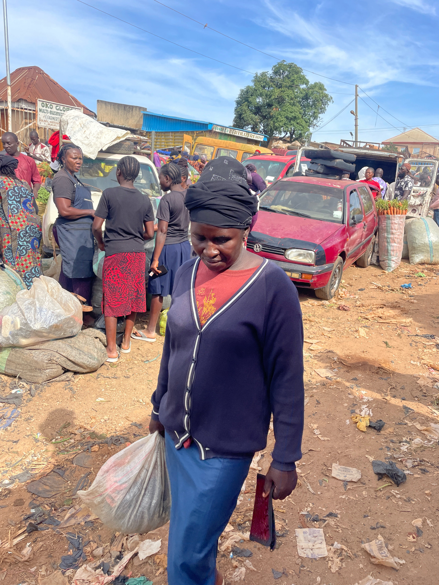 In Gura Top, Nigeria, a woman walks through a busy market carrying a large plastic bag. She is wearing a dark blue cardigan over an orange shirt and a blue skirt, with a black headscarf. Several other people are visible in the background, some engaged in what appears to be commerce, with goods such as produce and sacks of goods piled high. Cars and a yellow truck are also present, suggesting a lively marketplace environment. The ground is dusty and littered with debris, typical of an outdoor market setting.