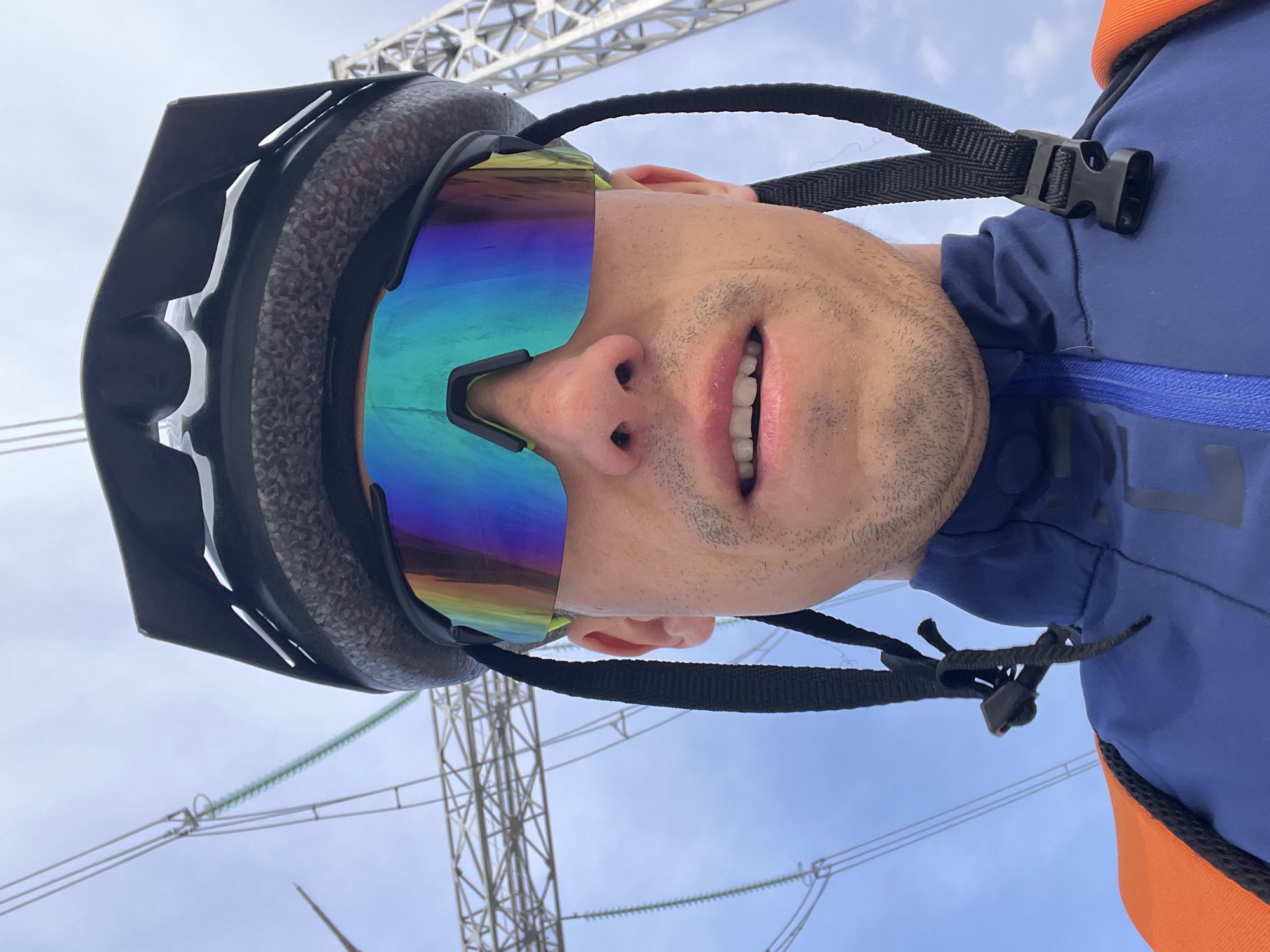 A man in a helmet and iridescent goggles looks directly at the camera from a low angle, suggesting he is lying on his back or looking up at the viewer. He is wearing a blue and orange jacket. In the background, there's a partially obscured power line tower. The photo may have been taken outdoors. There is insufficient information to determine the location of the city of Chaikovsky, Russia.