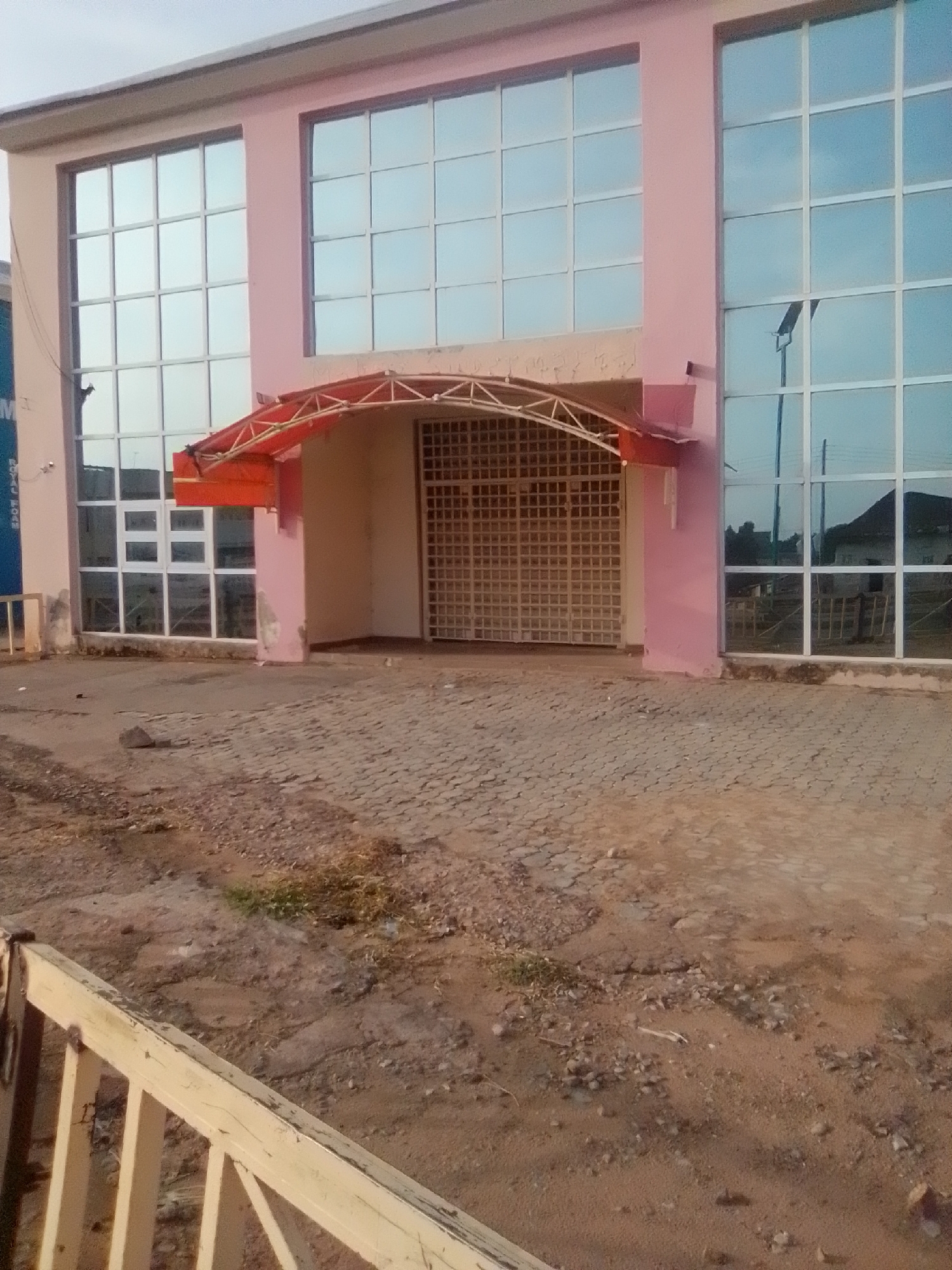 The image shows a pink building with large glass windows. There is a red awning over a metal gate. The ground in front of the building is paved with interlocking bricks and dirt. A yellow railing is visible in the foreground. The scene appears to be in an outdoor, semi-urban environment, likely in Pantame, Nigeria, given the context. No people are visible in the image.